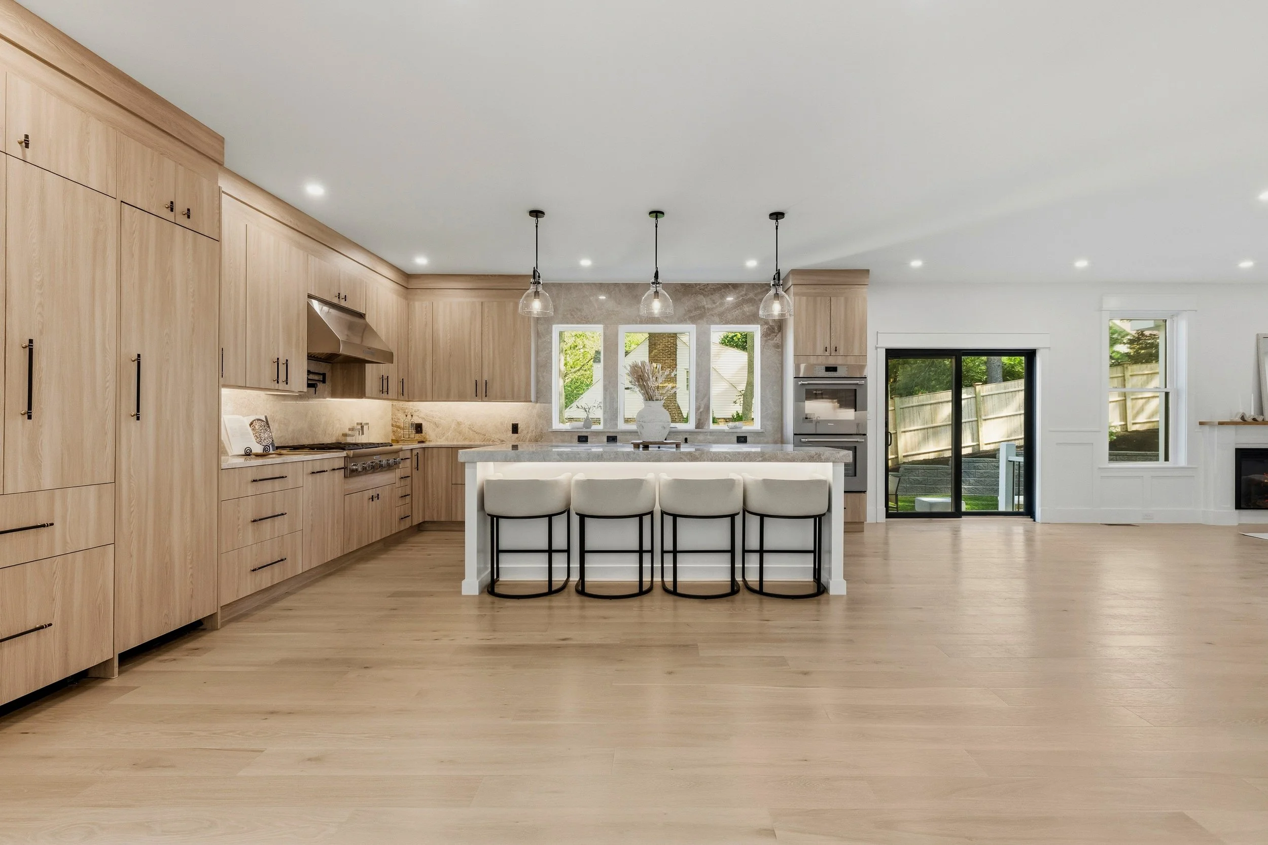 Modern kitchen with light wood cabinets, white island with four chairs, and large windows overlooking a backyard with a fence.
