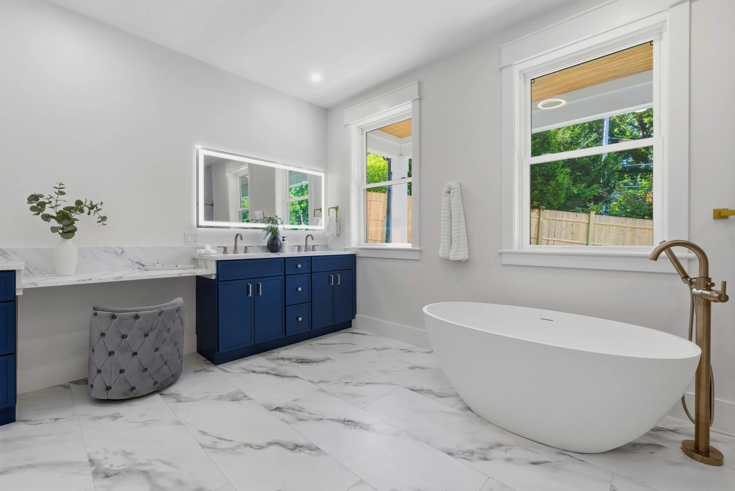 Modern bathroom with white walls, marble floors, a blue vanity with two sinks, a large mirror with LED lighting, a freestanding bathtub, and two windows showing green trees outside.