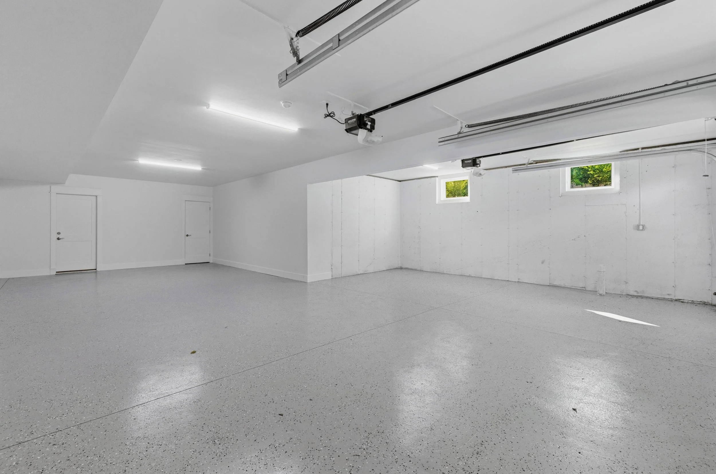 Empty garage with white walls, gray speckled epoxy floor, two small windows, and two garage door openers mounted on the ceiling.