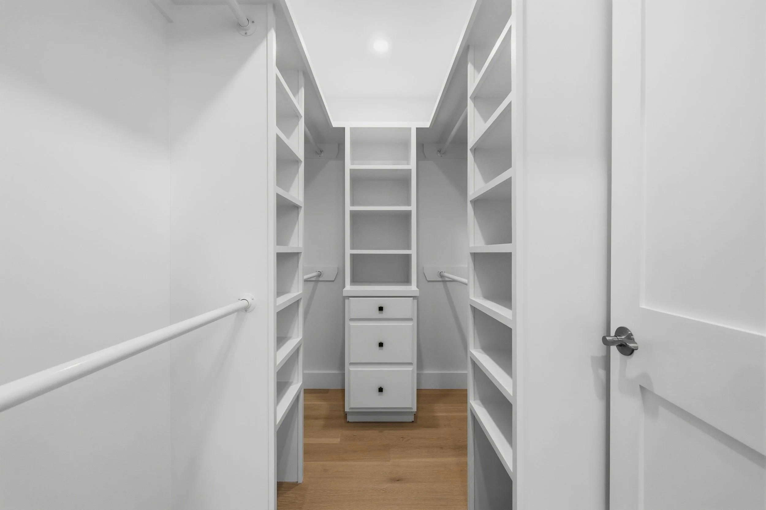 Empty white walk-in closet with built-in shelves and drawers, hardwood floors, and a door with a silver handle.
