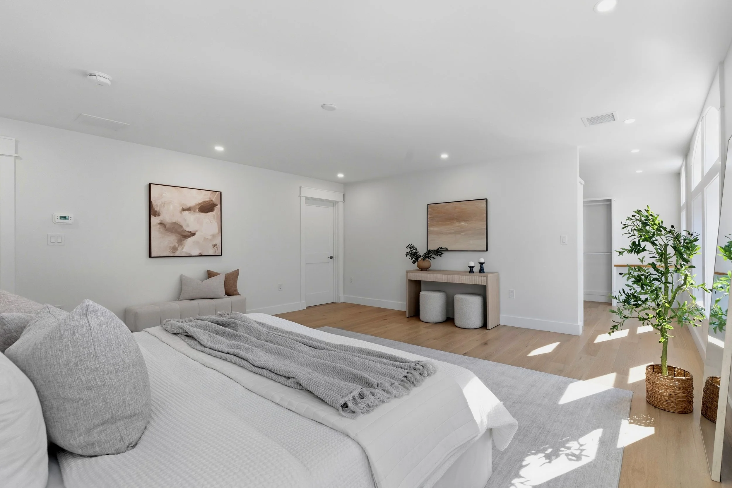 Bright modern bedroom with white bedding, gray pillows, abstract artwork on white walls, wooden flooring, a small desk with two stools, a potted plant, and natural light coming through large windows.