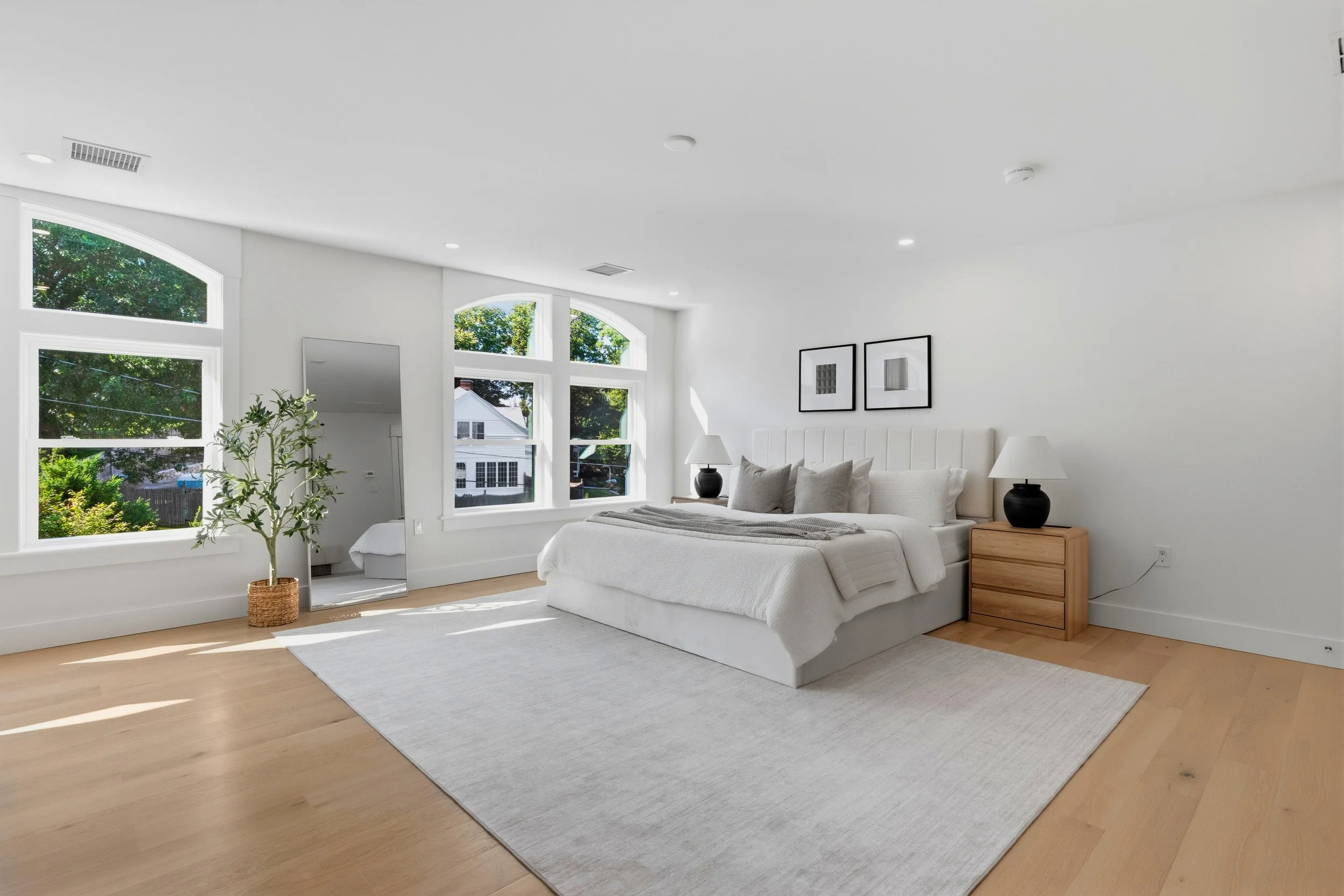 A bright, modern bedroom with large windows, a white bed with pillows and a blanket, two matching black lamps on wooden nightstands, framed minimalist art on the wall, a potted plant, and hardwood floors.