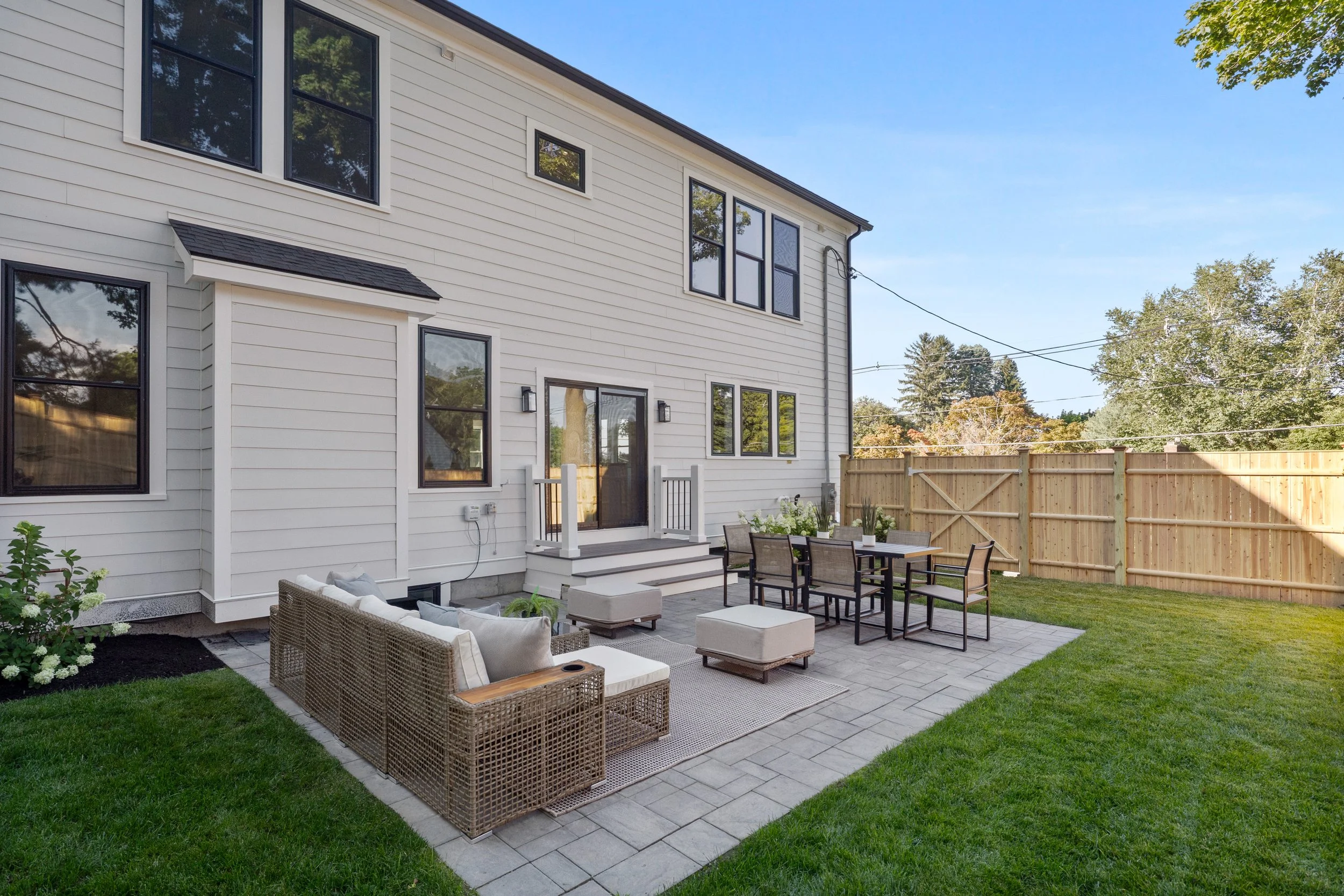A backyard patio area with outdoor furniture, including a wicker sofa and chairs, a dining table with chairs, and a small upholstered ottoman. The area is paved with gray bricks and is enclosed by a wooden fence. The house in the background has white