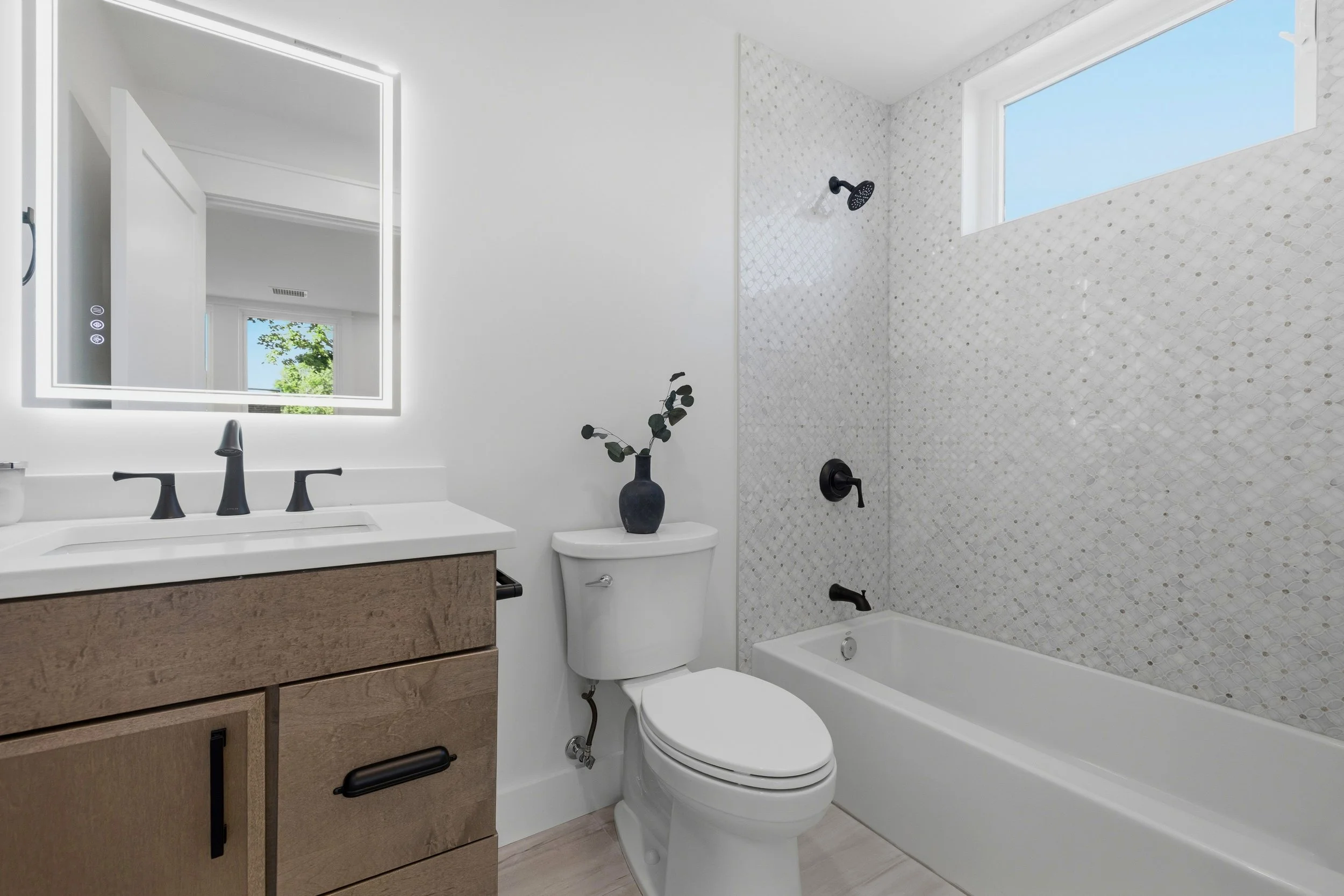 Modern bathroom with a white toilet, a wooden vanity with a white countertop, a large backlit mirror, and a bathtub with a showerhead. There is a small vase with a plant on the toilet tank, and a window with a clear blue sky outside.