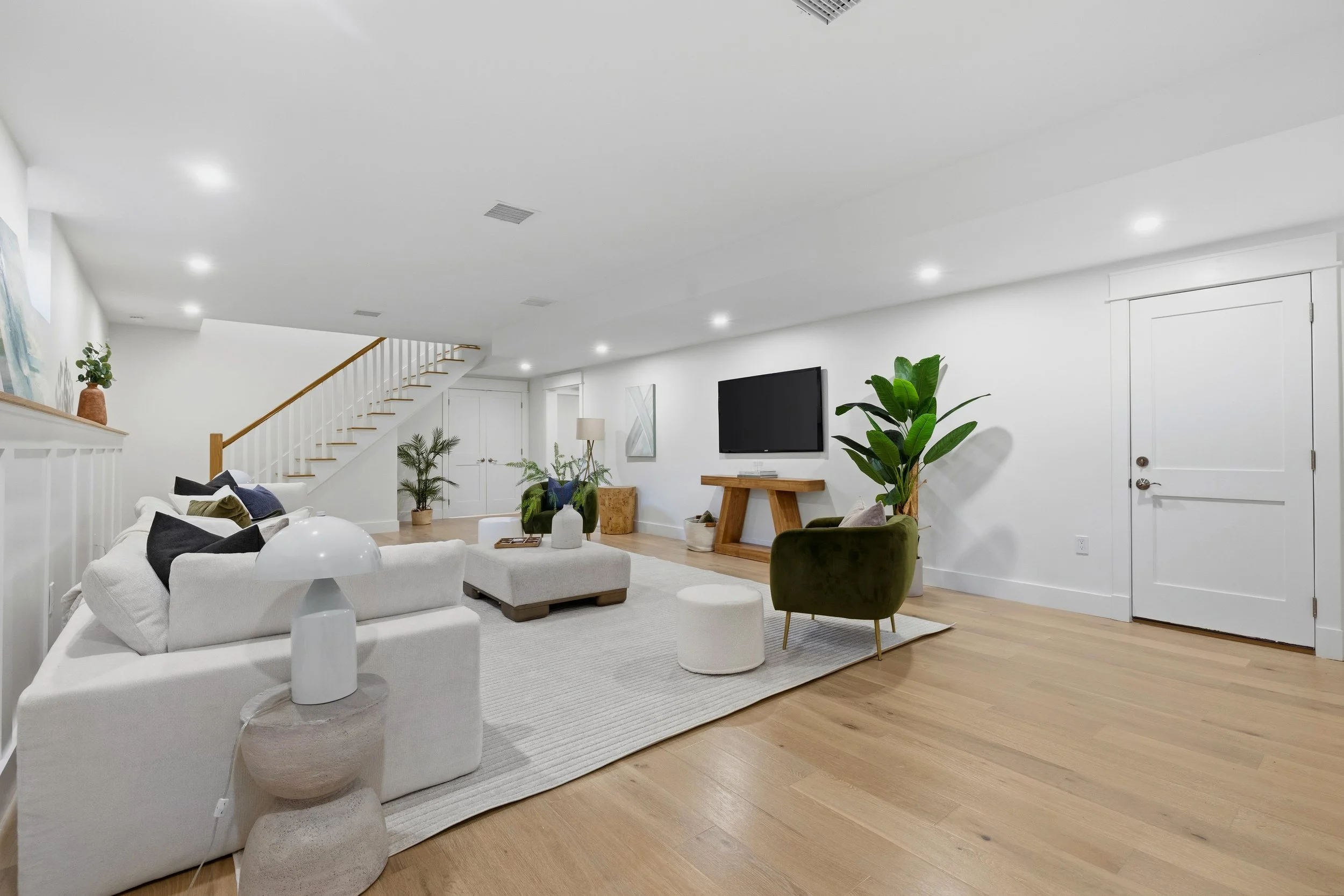 Living room with white walls and ceiling, hardwood floors, and modern furniture. There is a white sofa, a green velvet armchair, a wooden coffee table, and various plants. Mounted on the wall is a flat-screen TV. A staircase with white risers and woo