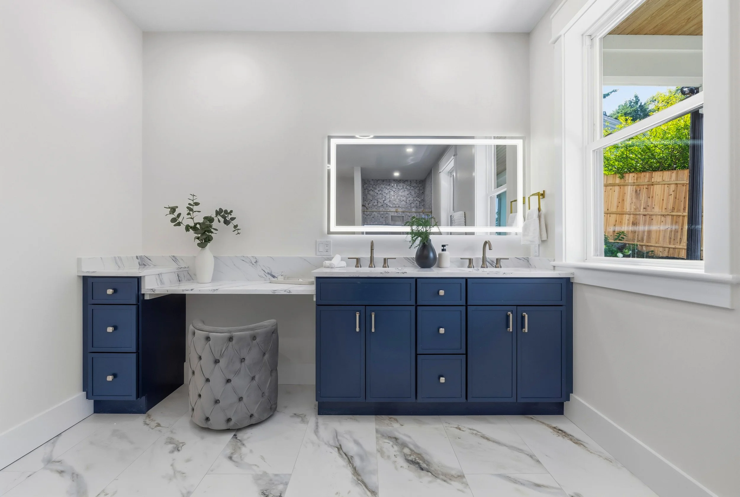 A bathroom vanity with blue cabinets, a white marble countertop, a large mirror with LED lighting, and a window showing a wooden fence and greenery outside.
