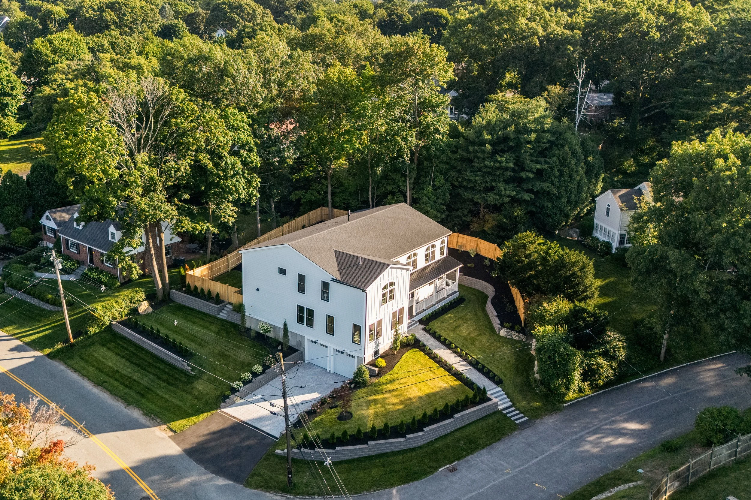 Aerial view of a residential neighborhood with large green trees, a white modern house with multiple levels, a well-maintained lawn, and surrounding streets.