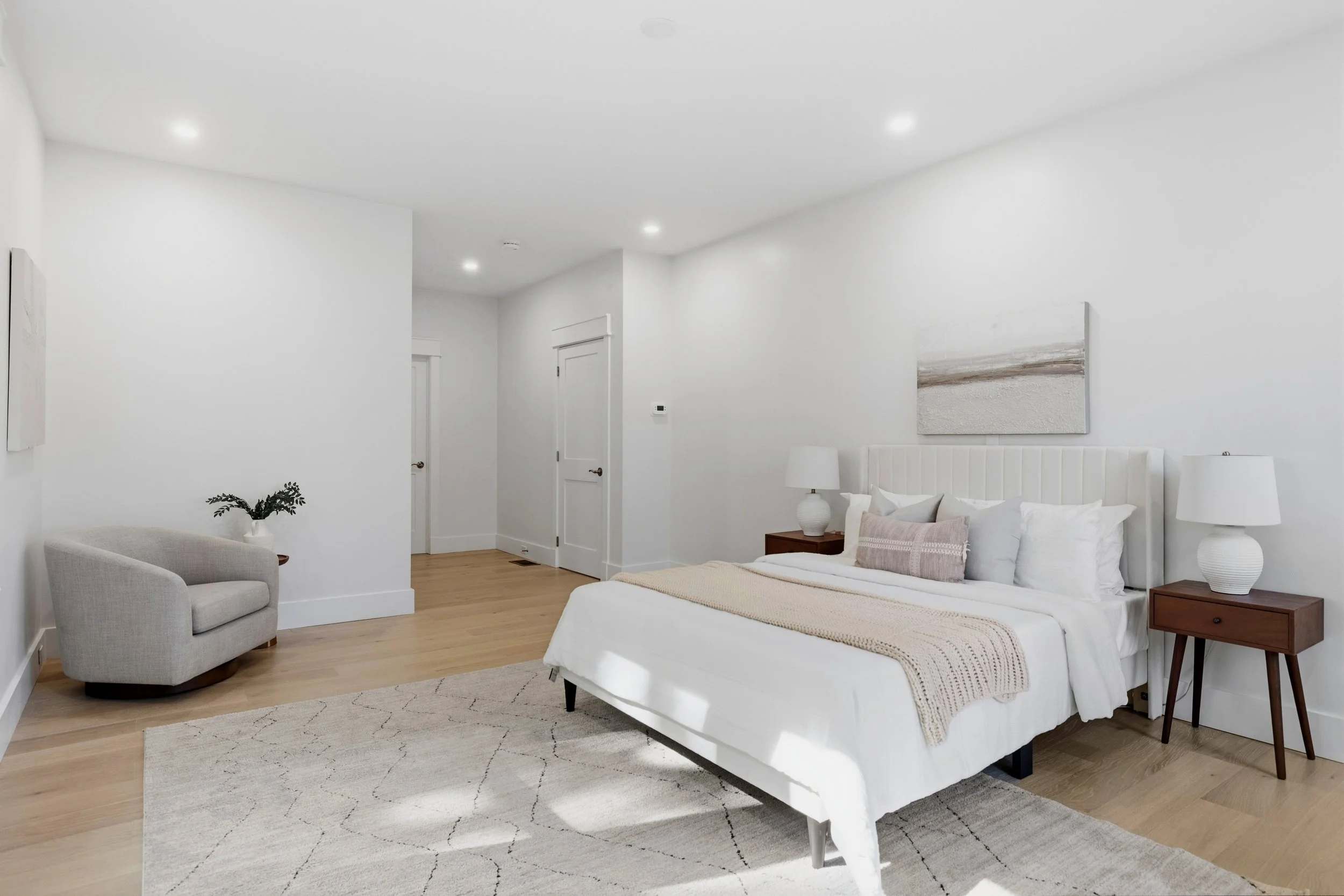 A bright, minimalist bedroom with white walls and a white bed with pillows. There are two white lamps on dark wood nightstands, a small gray armchair, and a painting of a beach scene above the bed. A white rug with a geometric pattern is on a light w