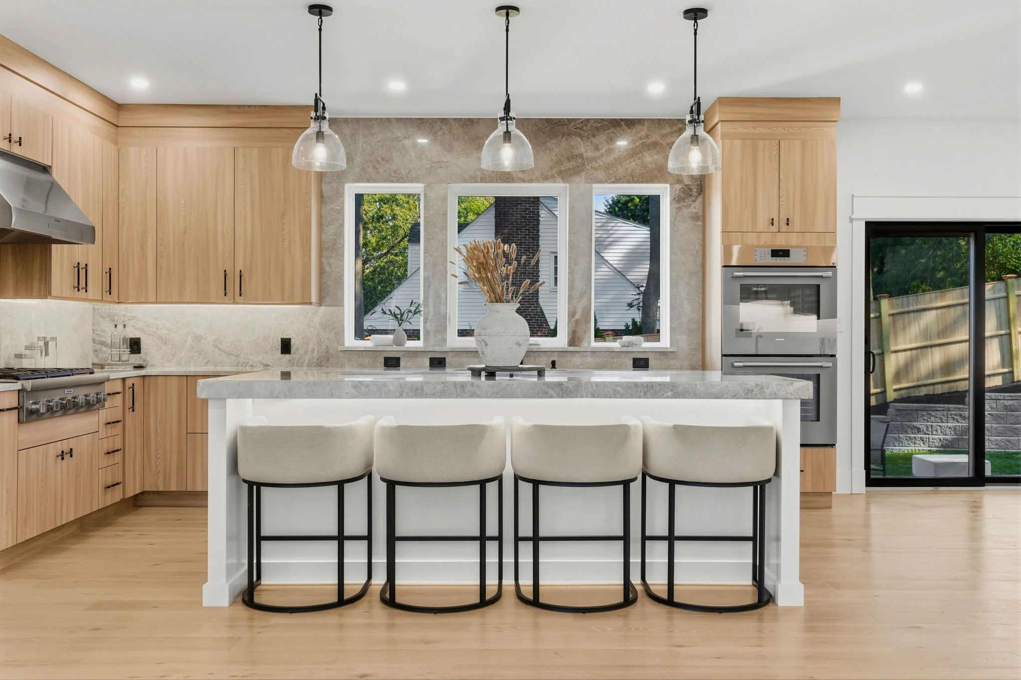 Modern kitchen with light wood cabinets, a marble island with four white chairs, pendant lights hanging from the ceiling, a large window, and a sliding glass door leading outside.