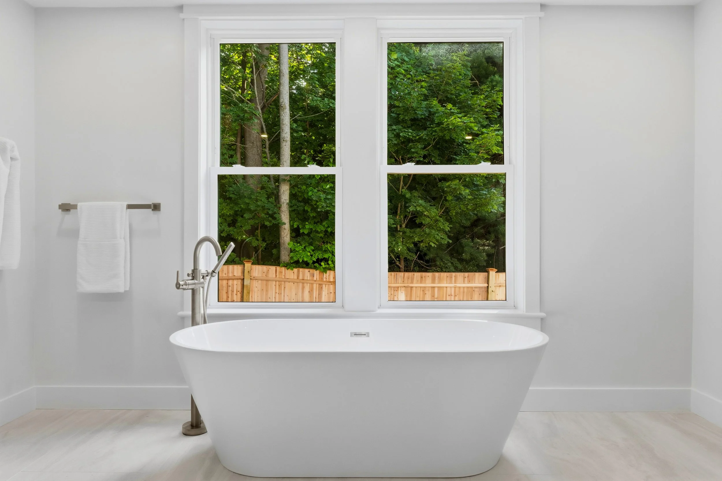 White bathtub in front of a large window with a view of green trees and a wooden fence outside.