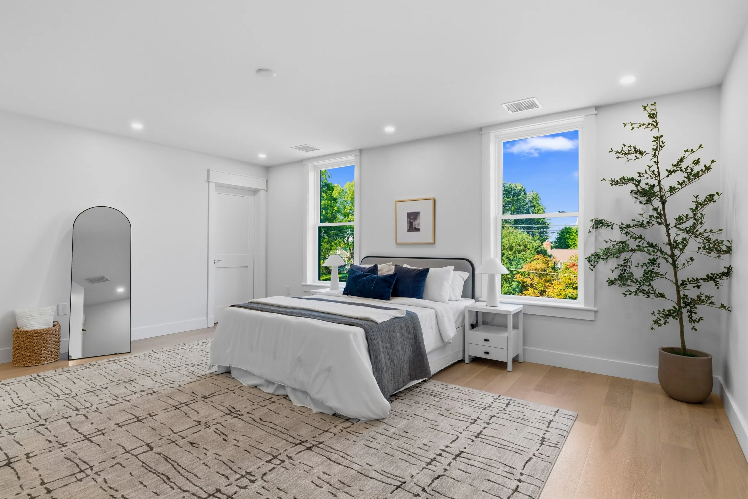 Bright bedroom with white walls and a large window showing green trees and blue sky, a bed with white bedding and dark blue pillows, a white nightstand, a potted plant, and a large mirror.