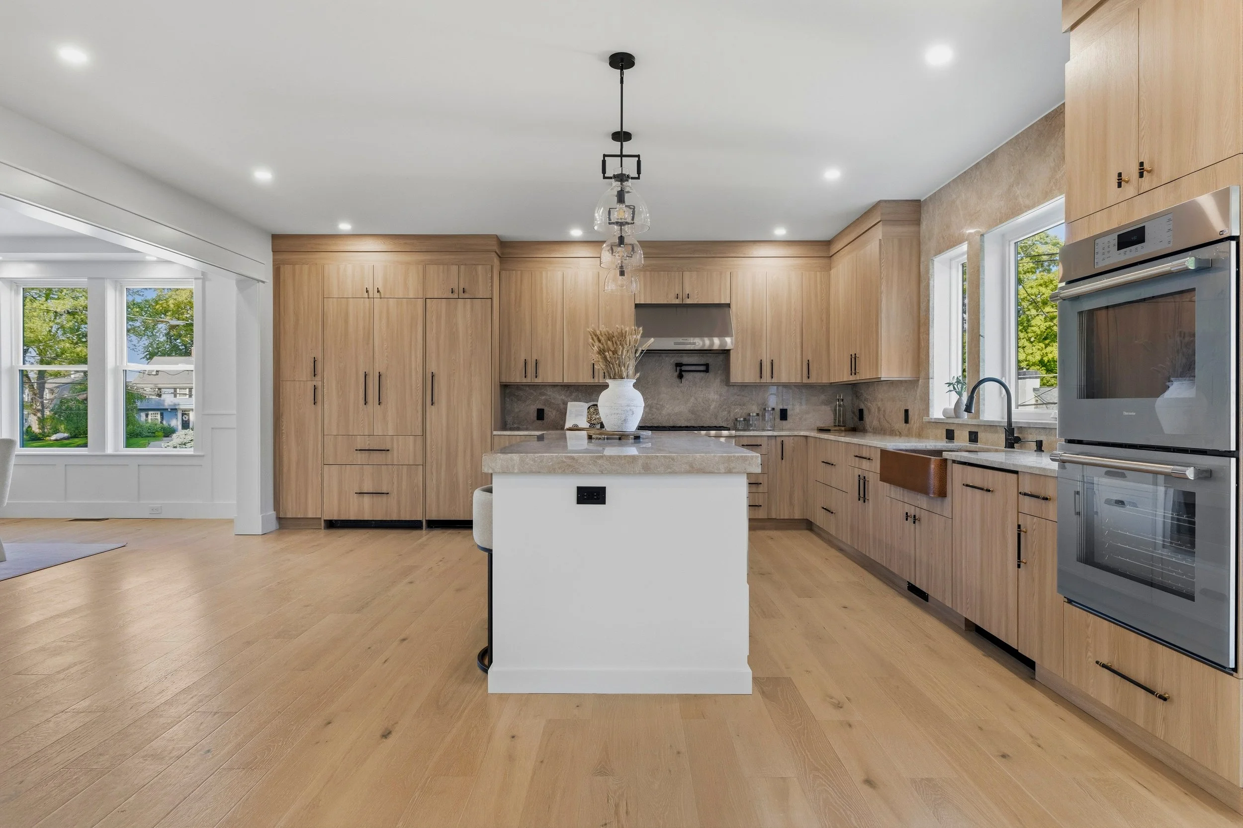 Modern kitchen with wooden cabinets, a kitchen island with a white base, a vase with dried flowers, stainless steel appliances, and large windows with views of green trees outside.