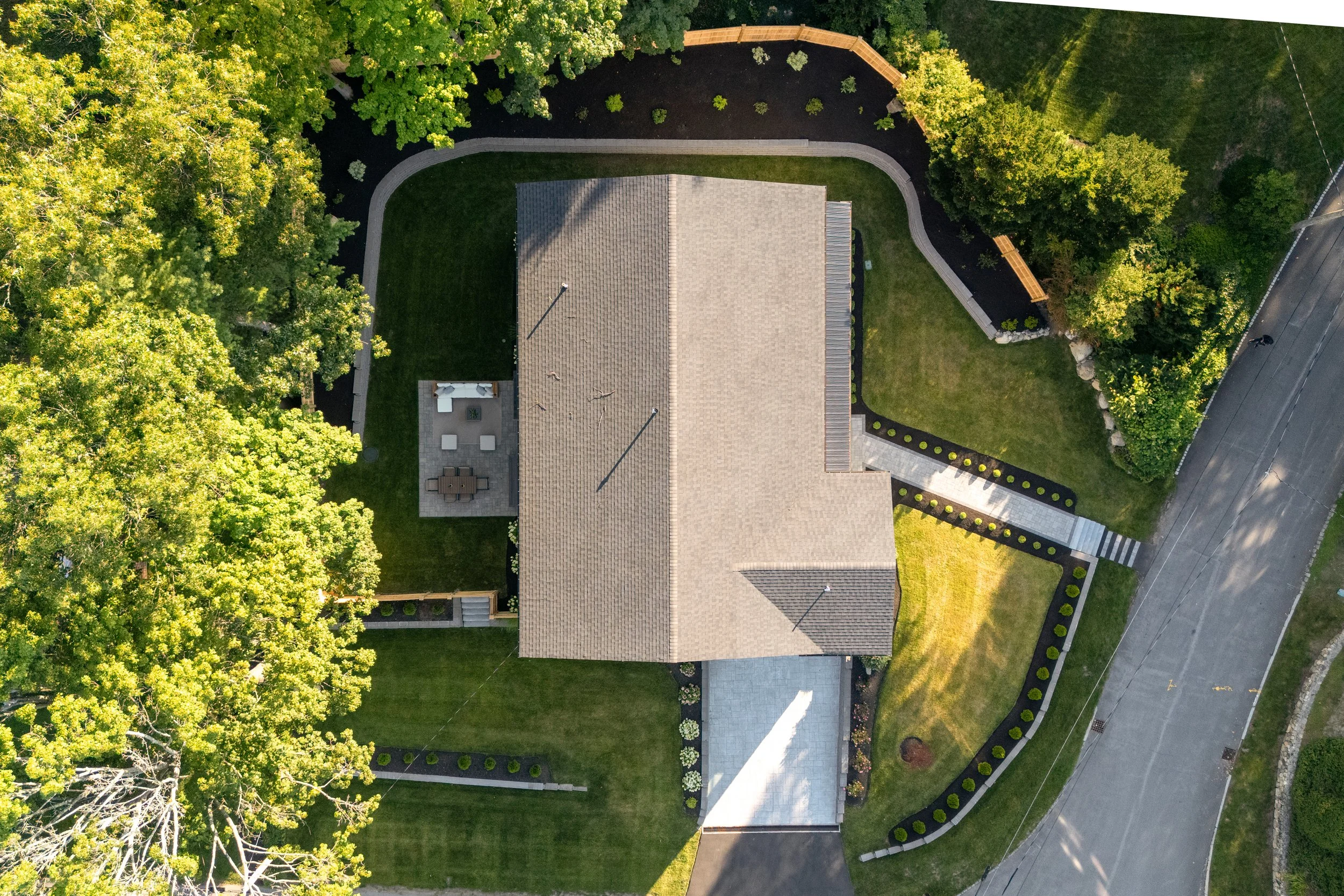 An aerial view of a house surrounded by a well-maintained yard, trees, and a curved driveway in a suburban neighborhood.