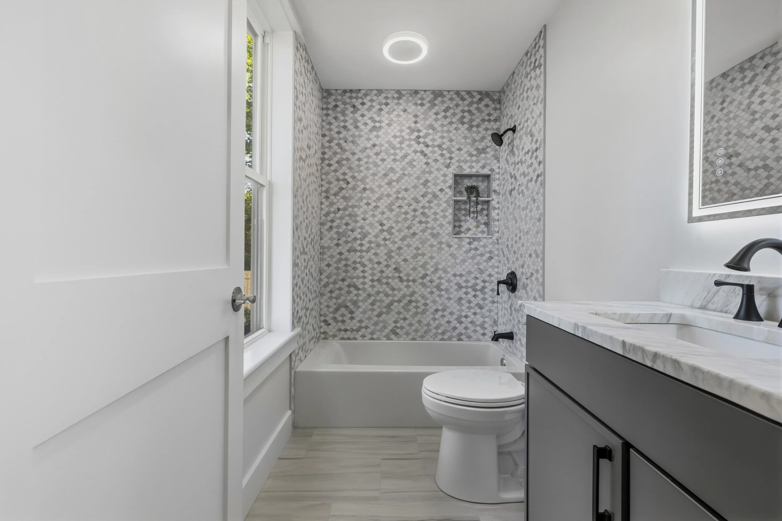 Modern bathroom with white walls, a large window, a walk-in shower with gray and white hexagonal tiles, a built-in niche with a plant, a white toilet, and a gray vanity with a marble countertop and black fixtures.