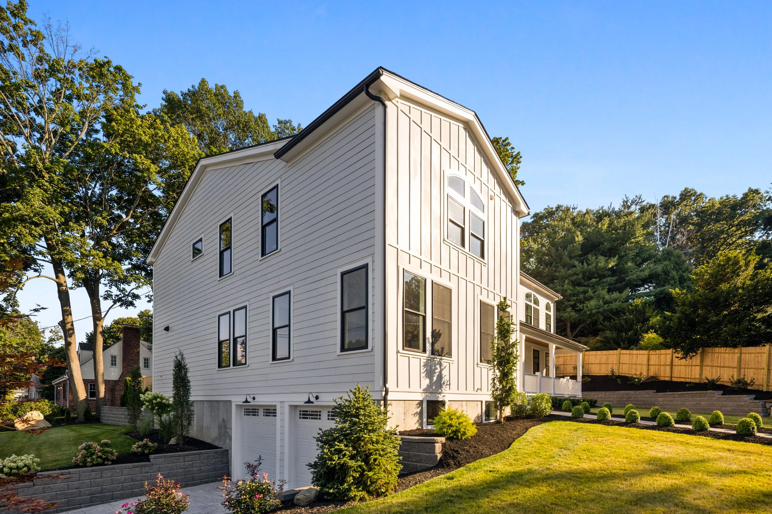 A white multi-story house with tall, narrow windows on a sunny day, surrounded by a landscaped yard with green grass, small bushes, and trees, with a blue sky and wooded area in the background.