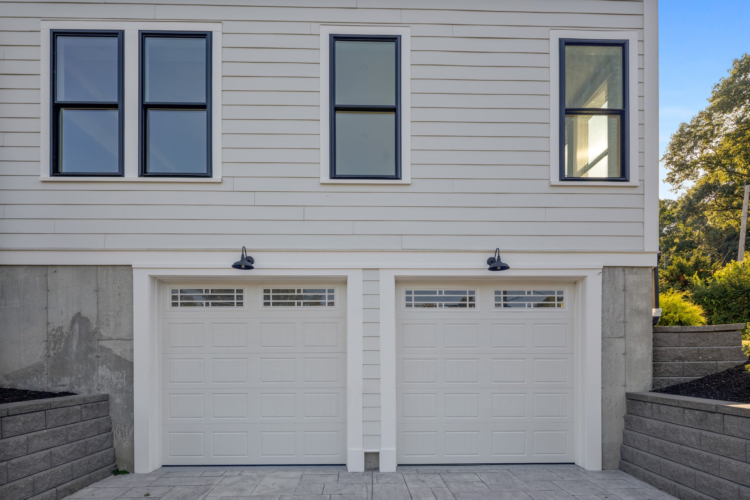Front of a house with a white double garage door, white siding, three windows above, two wall-mounted black outdoor lights, and a concrete and stone yard with stone walls on each side.