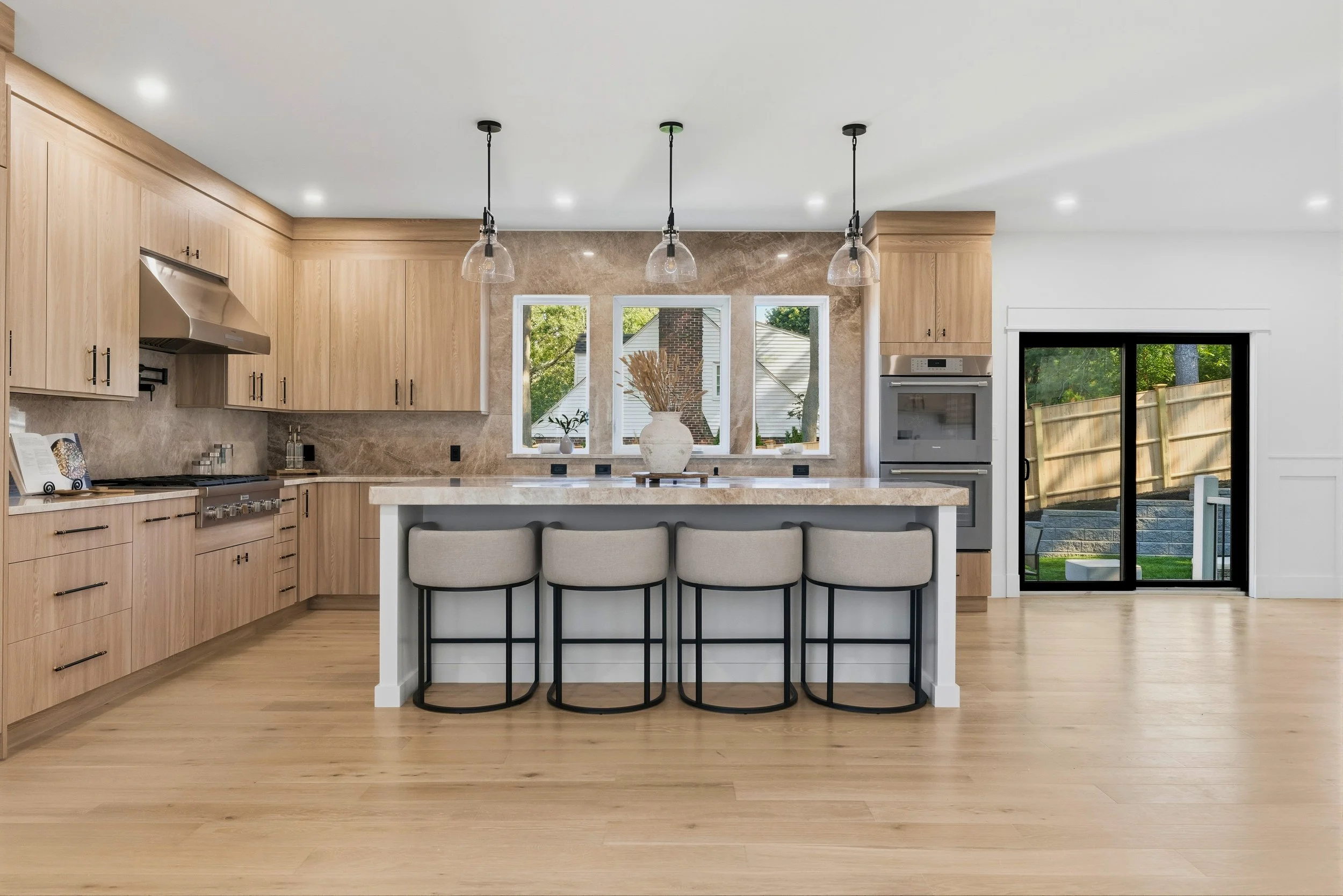 Modern kitchen with light wood cabinets, marble countertops, and a kitchen island with four beige barstools. Three pendant lights hang above the island. There are three windows behind the sink with outdoor views, including trees and neighboring house