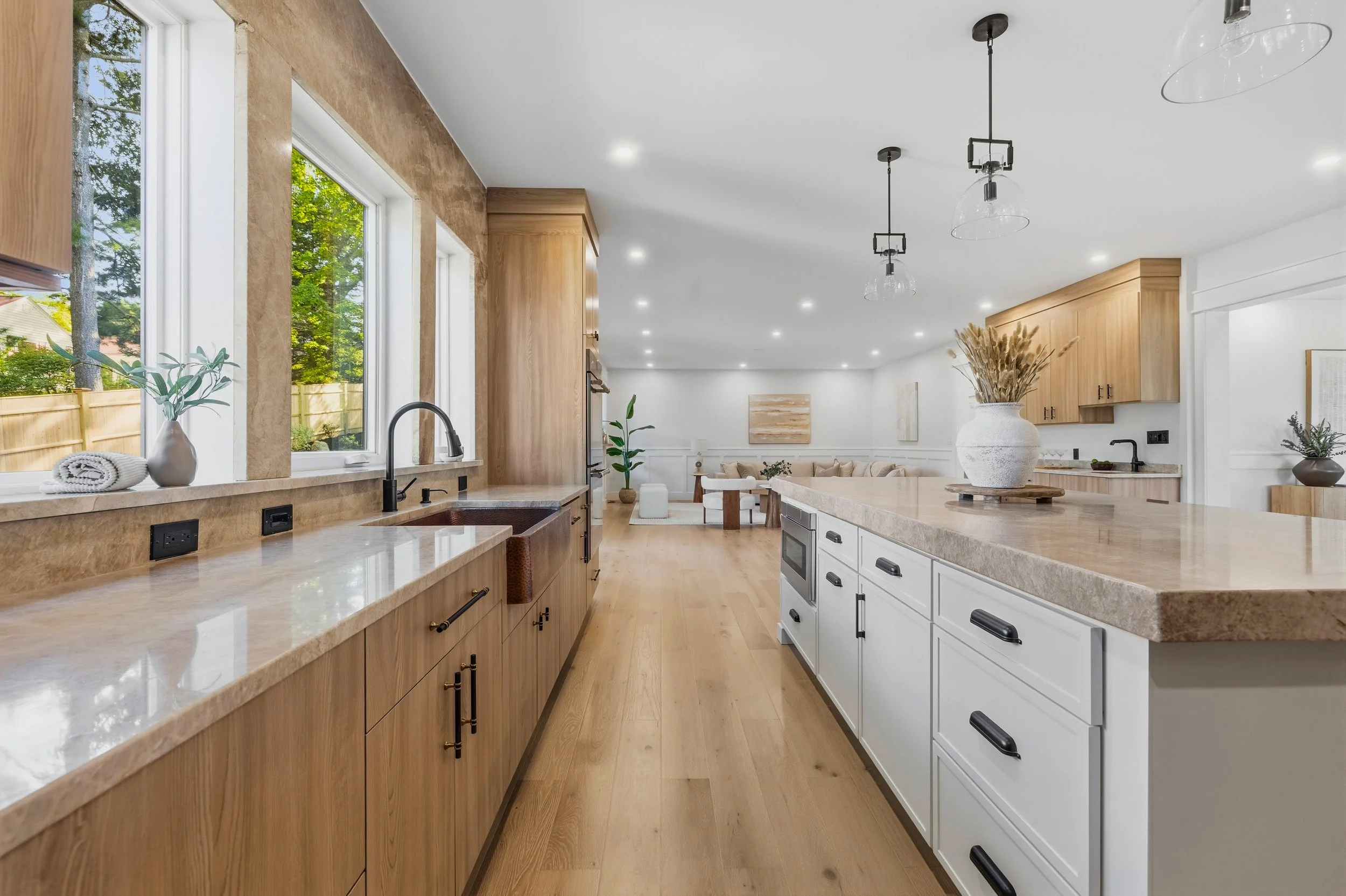 Modern kitchen with natural wood cabinets, white island, large windows, and pendant lighting, open to a living space with beige sofa and artwork.