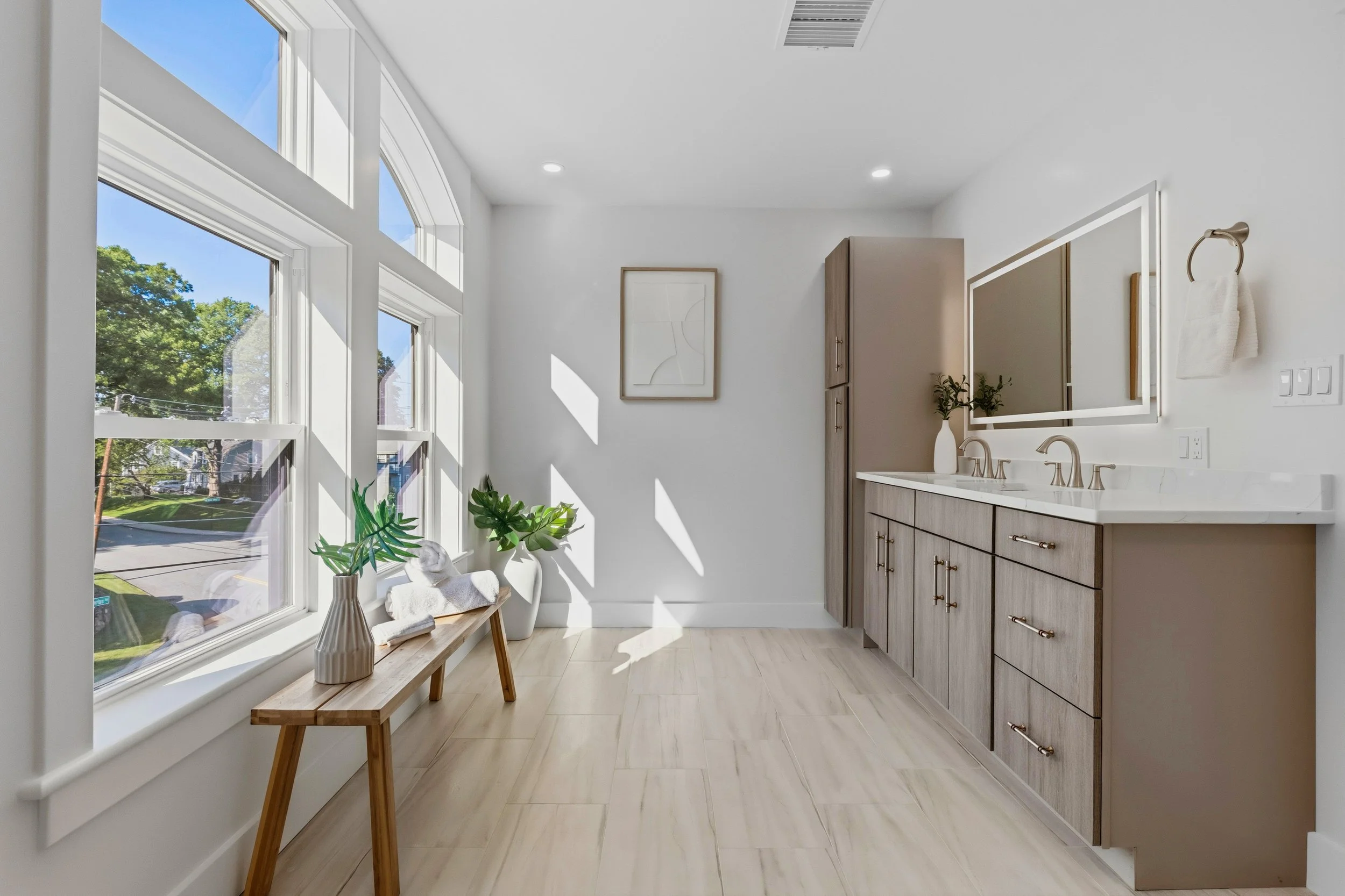 Bright bathroom with large windows, white walls, a double vanity with beige cabinets, a mirror, and a small bench with decorative vases and plants.