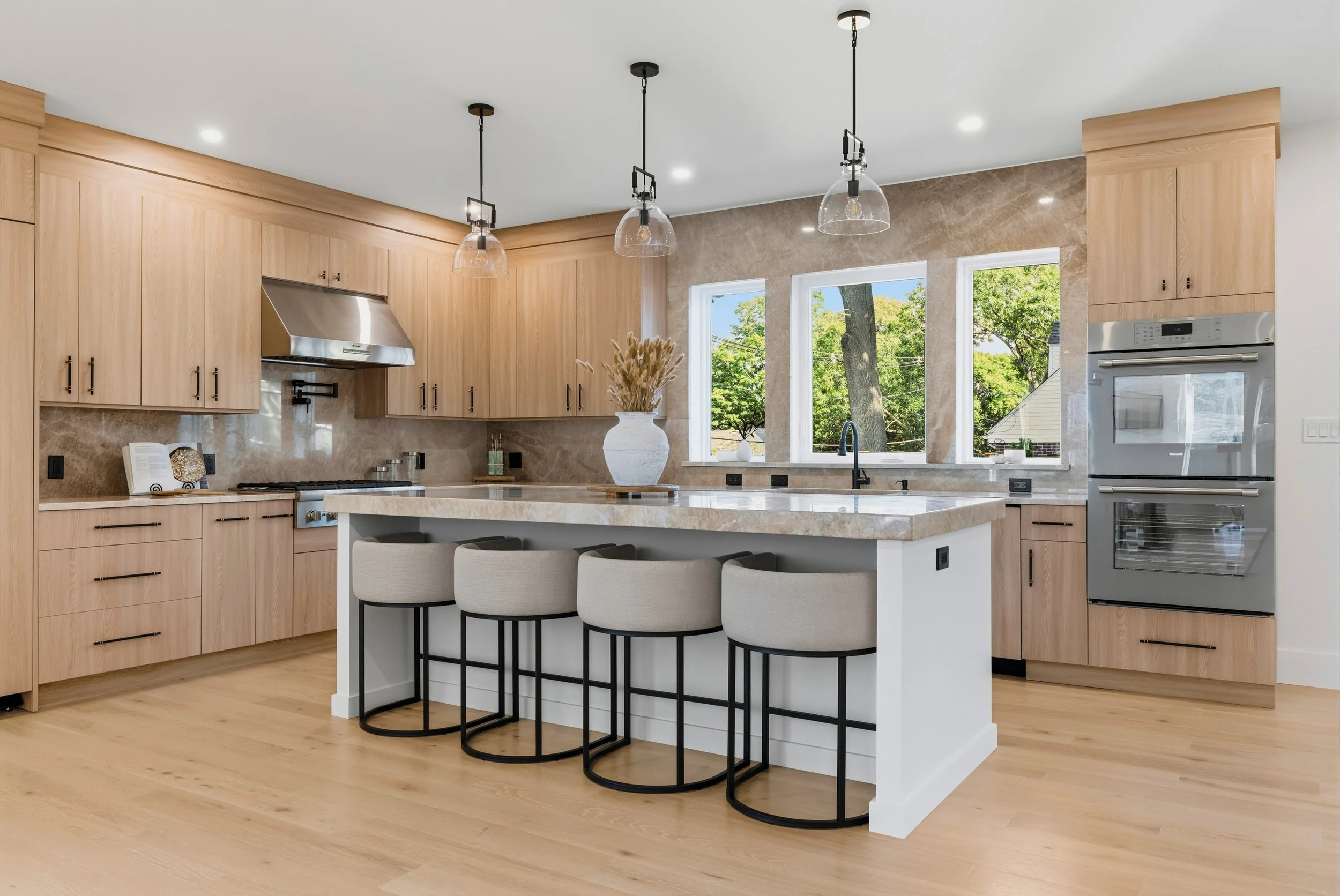 Modern kitchen with light wood cabinets, a large white island with seating, three pendant lights, large windows showing trees outside, and stainless steel appliances.