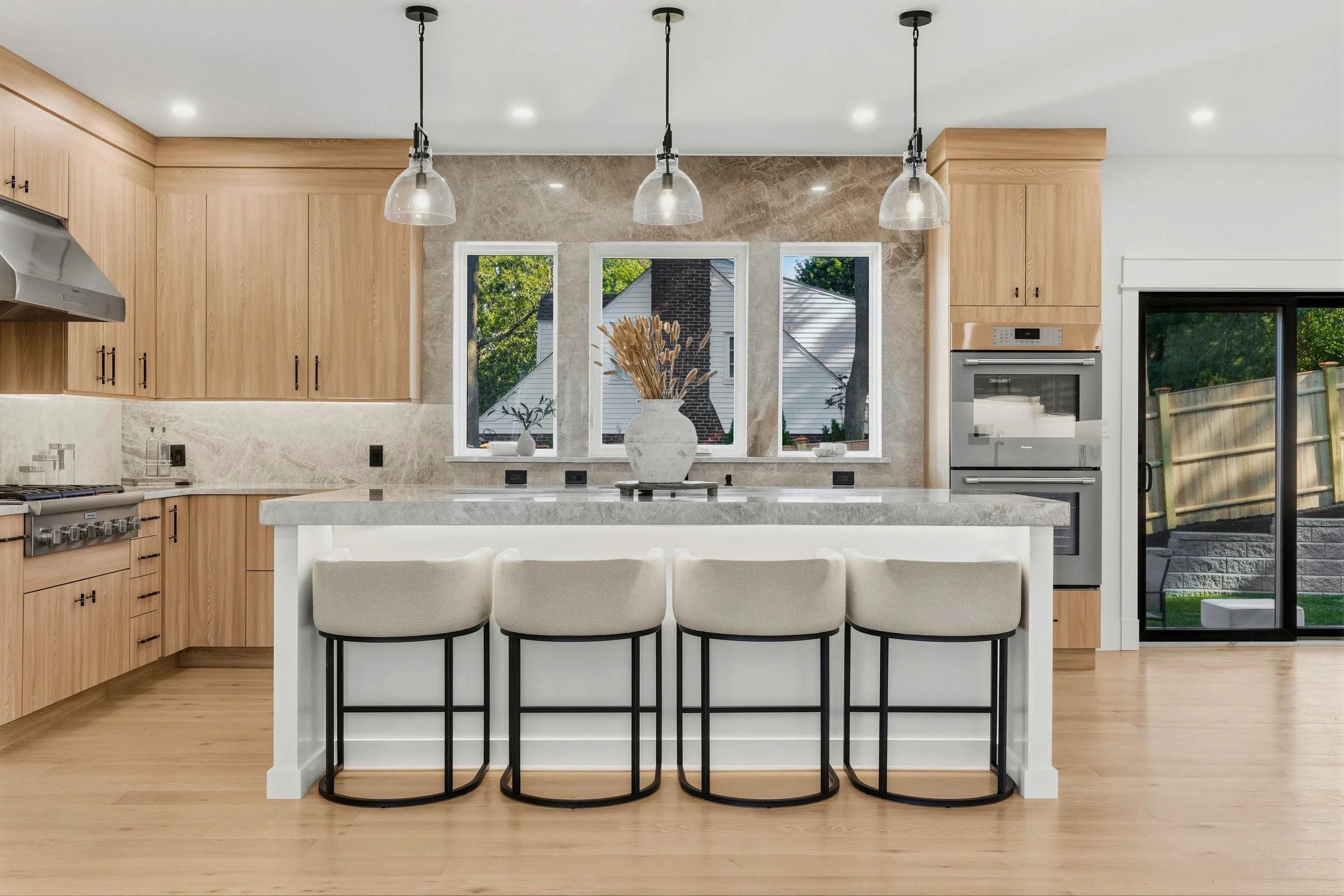 Modern kitchen with wooden cabinets, white island with four beige chairs, large window, and glass pendant lights hanging from the ceiling.