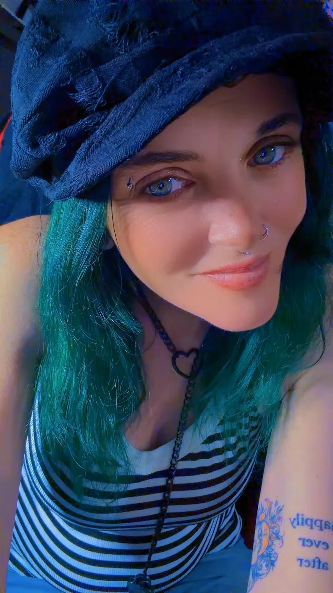 A woman with blue-green hair, wearing a black cap, striped tank top, and multiple facial piercings, smiling at the camera.