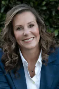 A woman with wavy brown hair smiling, wearing a white shirt and a dark blue blazer, outdoors with green foliage in the background.