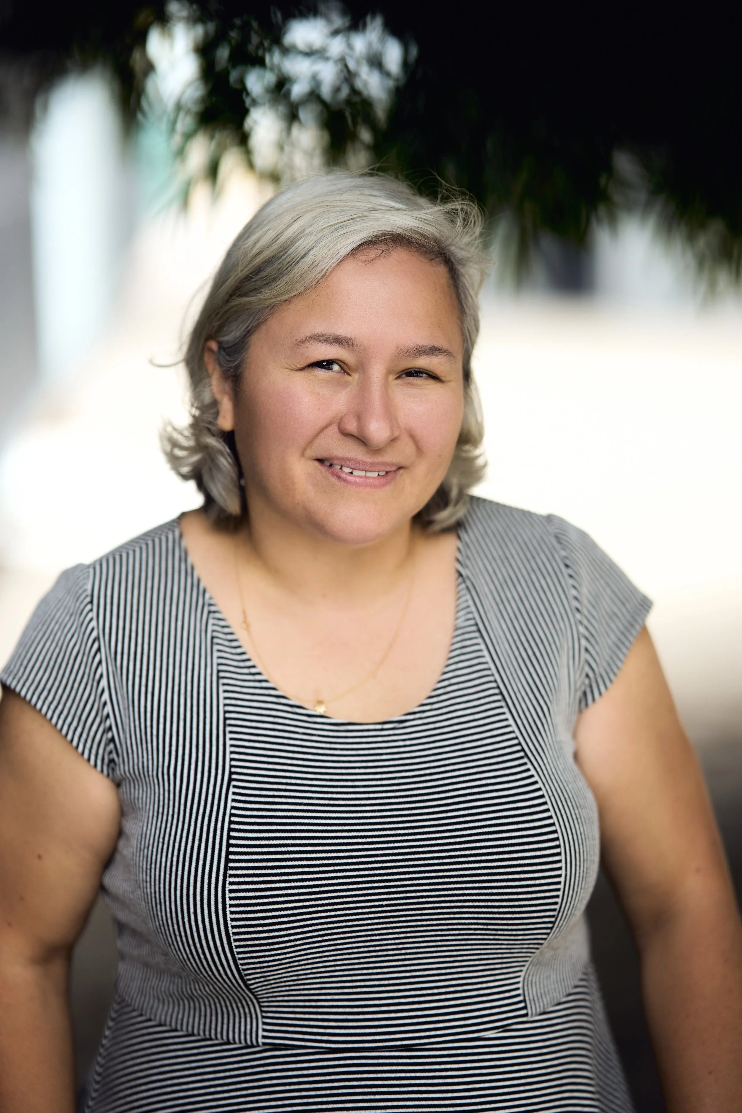 A woman with short gray hair, wearing a striped gray and white top, smiling outdoors with blurred trees in the background.