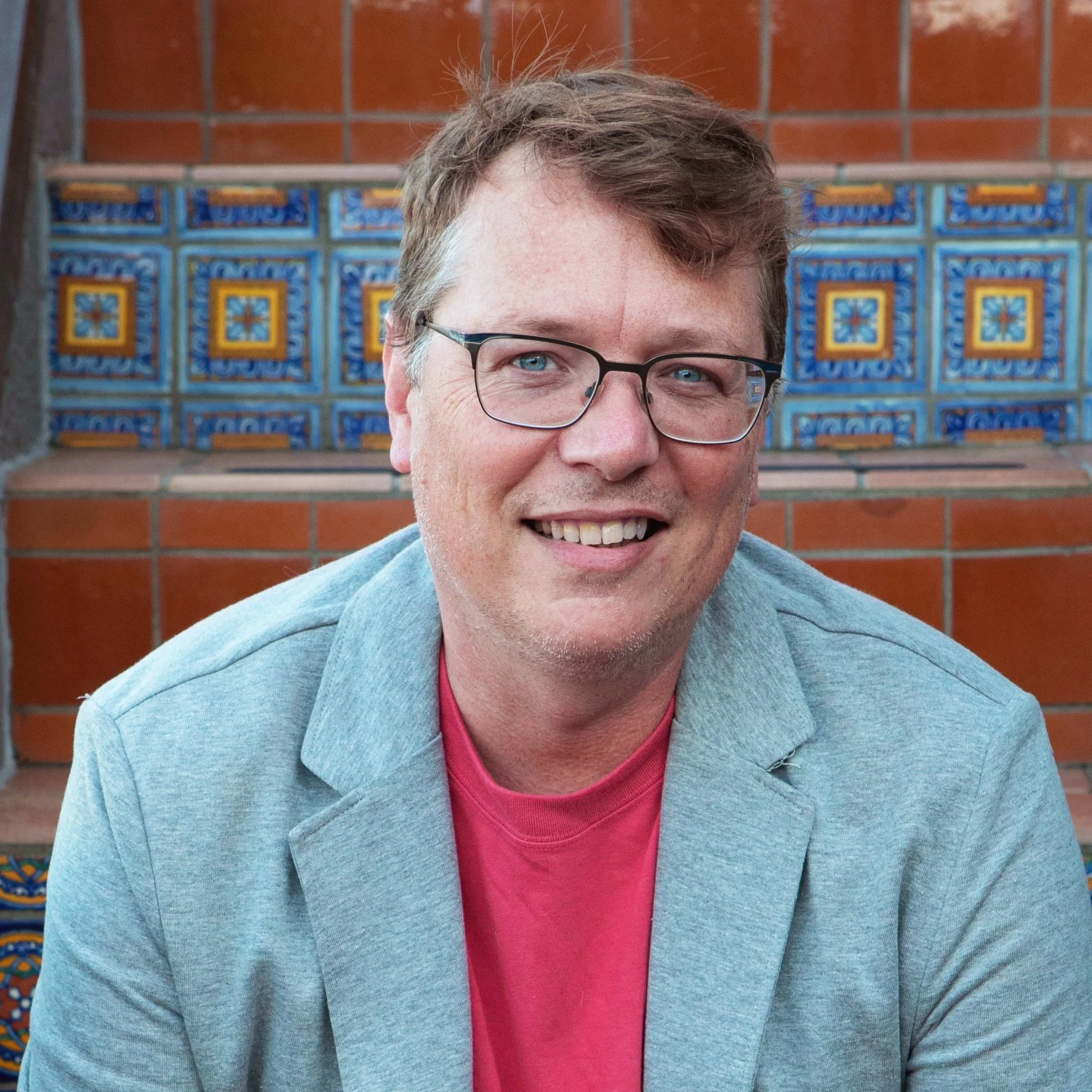 Partial close-up of a smiling man with glasses, wearing a blue blazer and red shirt, standing in front of a colorful tiled wall.