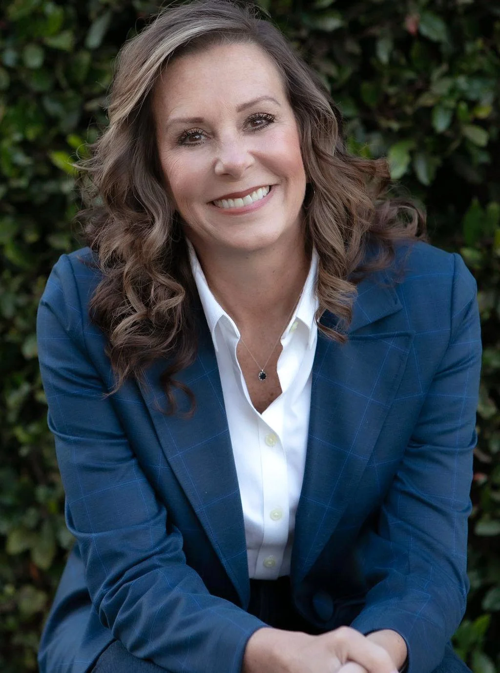 A woman with shoulder-length curly brown hair, smiling, wearing a blue blazer over a white shirt, with a black pendant necklace, sitting outdoors in front of green foliage.