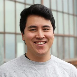 A young man with dark hair smiling, standing in front of a modern building with glass windows.