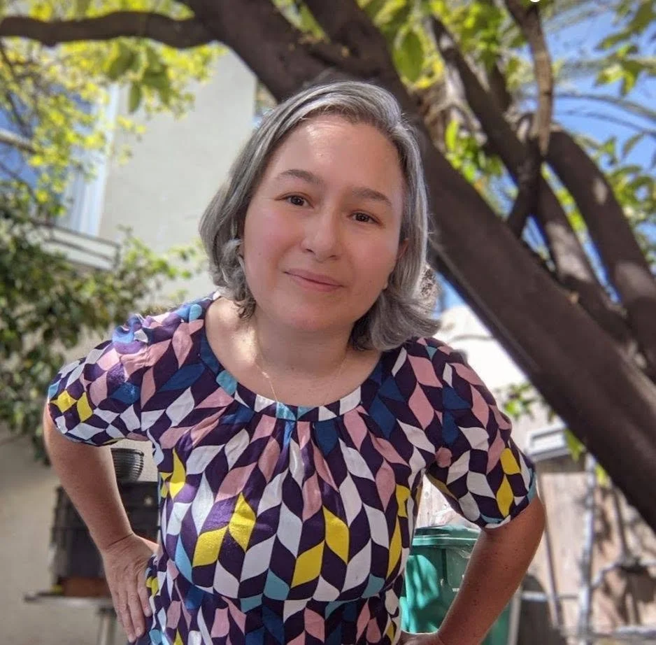 A woman with short gray hair and a friendly smile standing outdoors in a garden, wearing a colorful geometric patterned blouse, with trees and a house in the background.