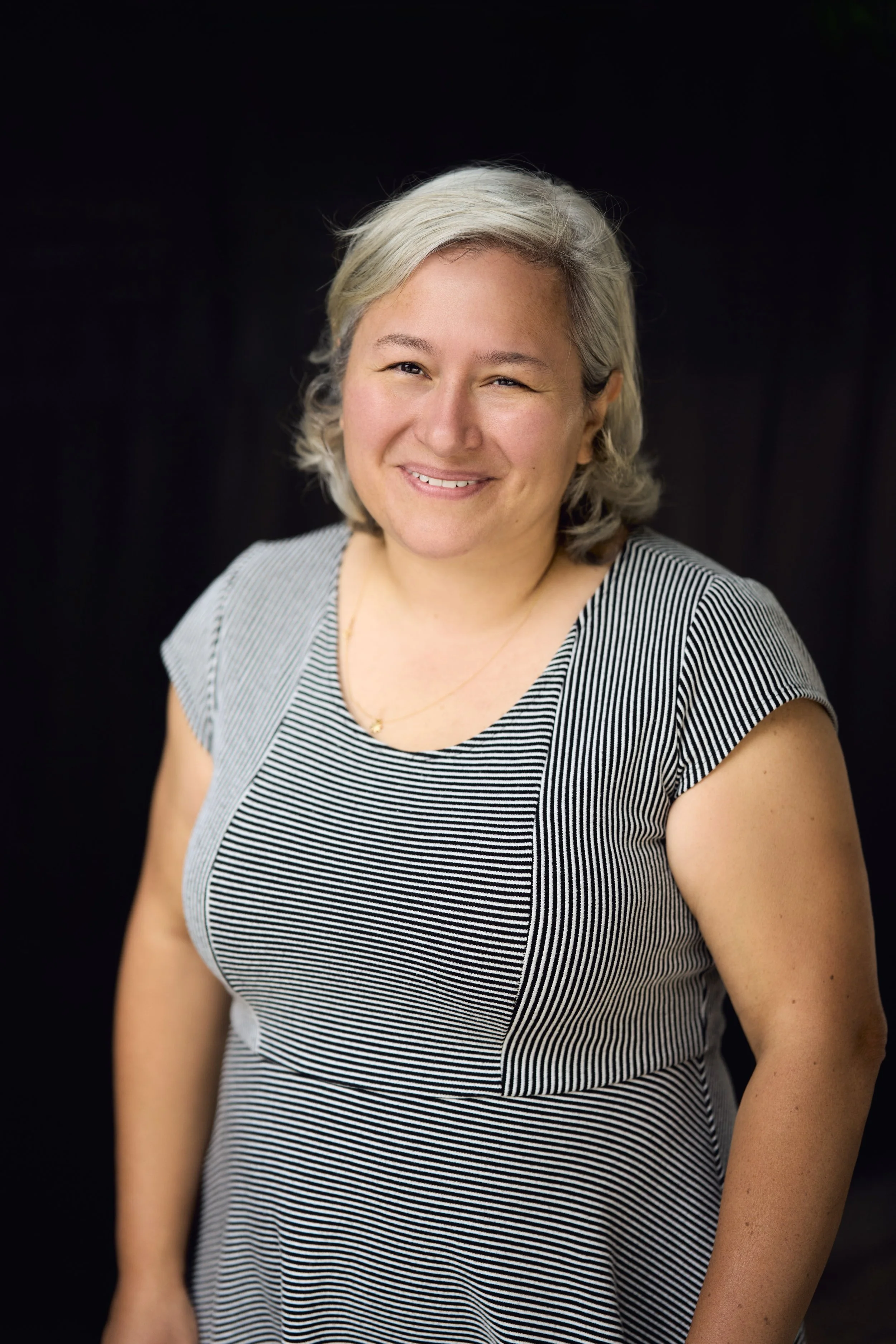 A woman with short blonde hair, smiling and wearing a black and white striped shirt, standing against a black background.