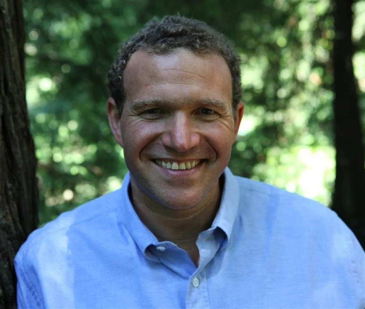 A man smiling outdoors with green trees in the background, wearing a light blue button-up shirt.