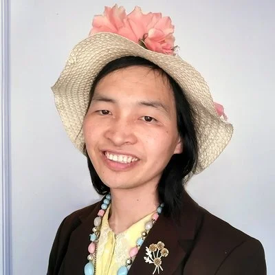 Person wearing a wide-brimmed straw hat with pink flowers, a brown blazer, and a colorful necklace, smiling against a light background.