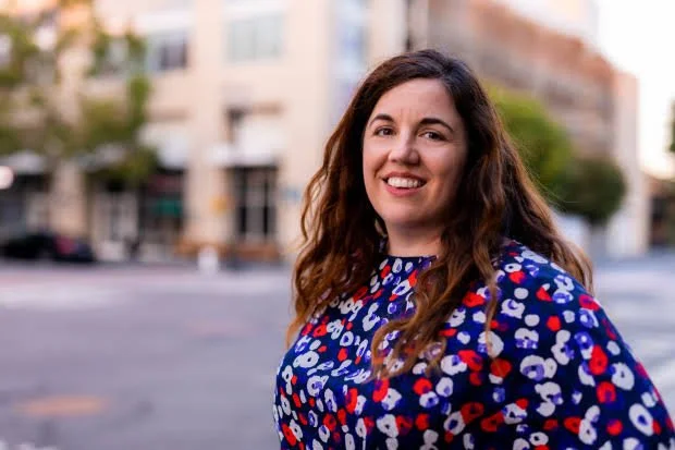 Woman with long curly hair smiling outdoors in front of an urban building, wearing a patterned blue, red, and white blouse.