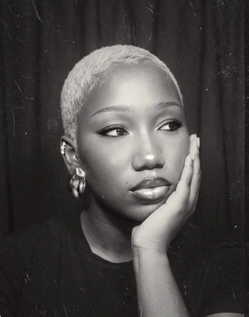 Black and white portrait of a woman with short platinum blonde hair, wearing earrings, resting her face on her hand and gazing thoughtfully to the side.