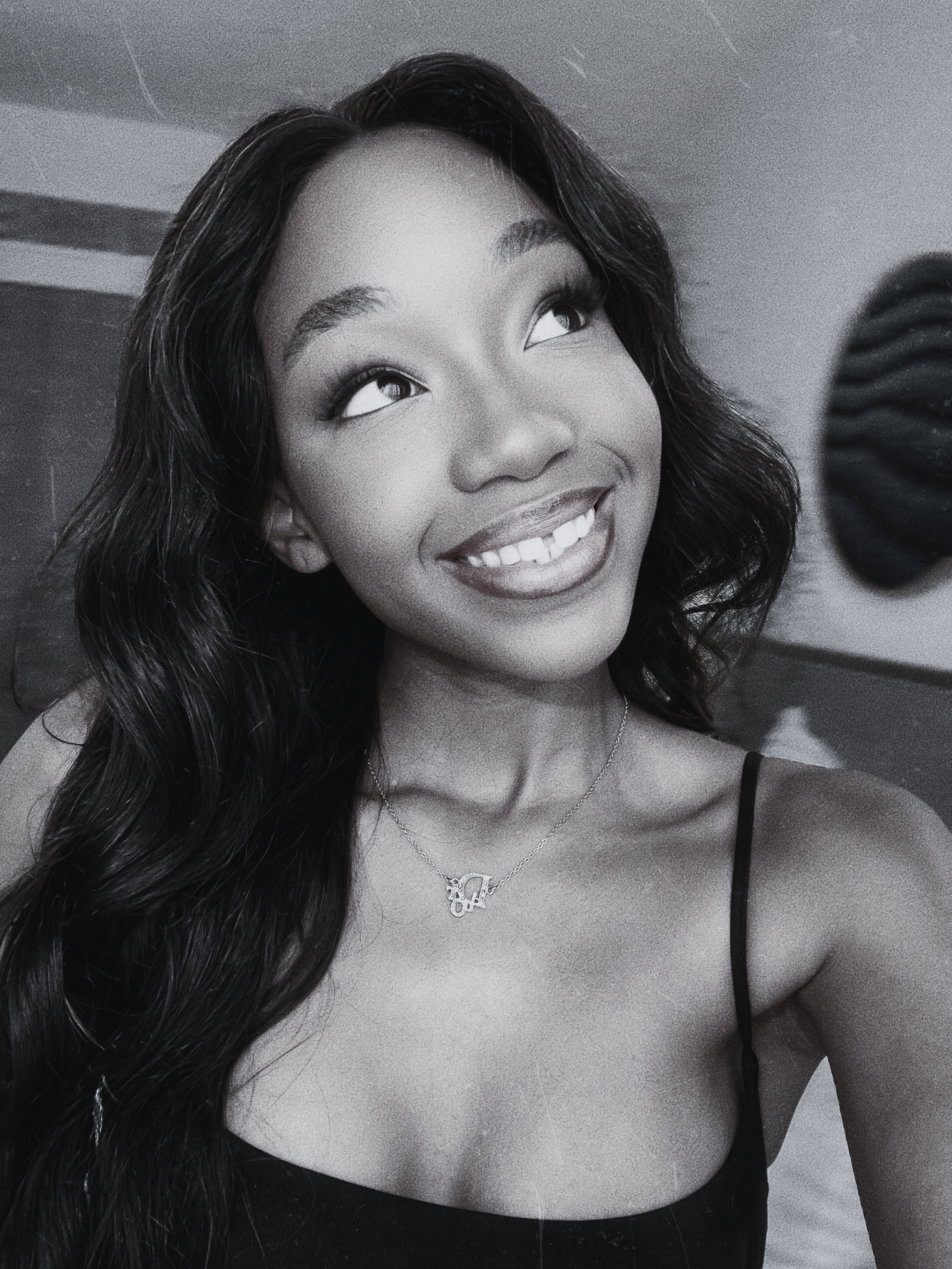 Close-up of a young woman with long dark wavy hair, smiling and looking upwards, wearing a necklace with a pendant and a black top.