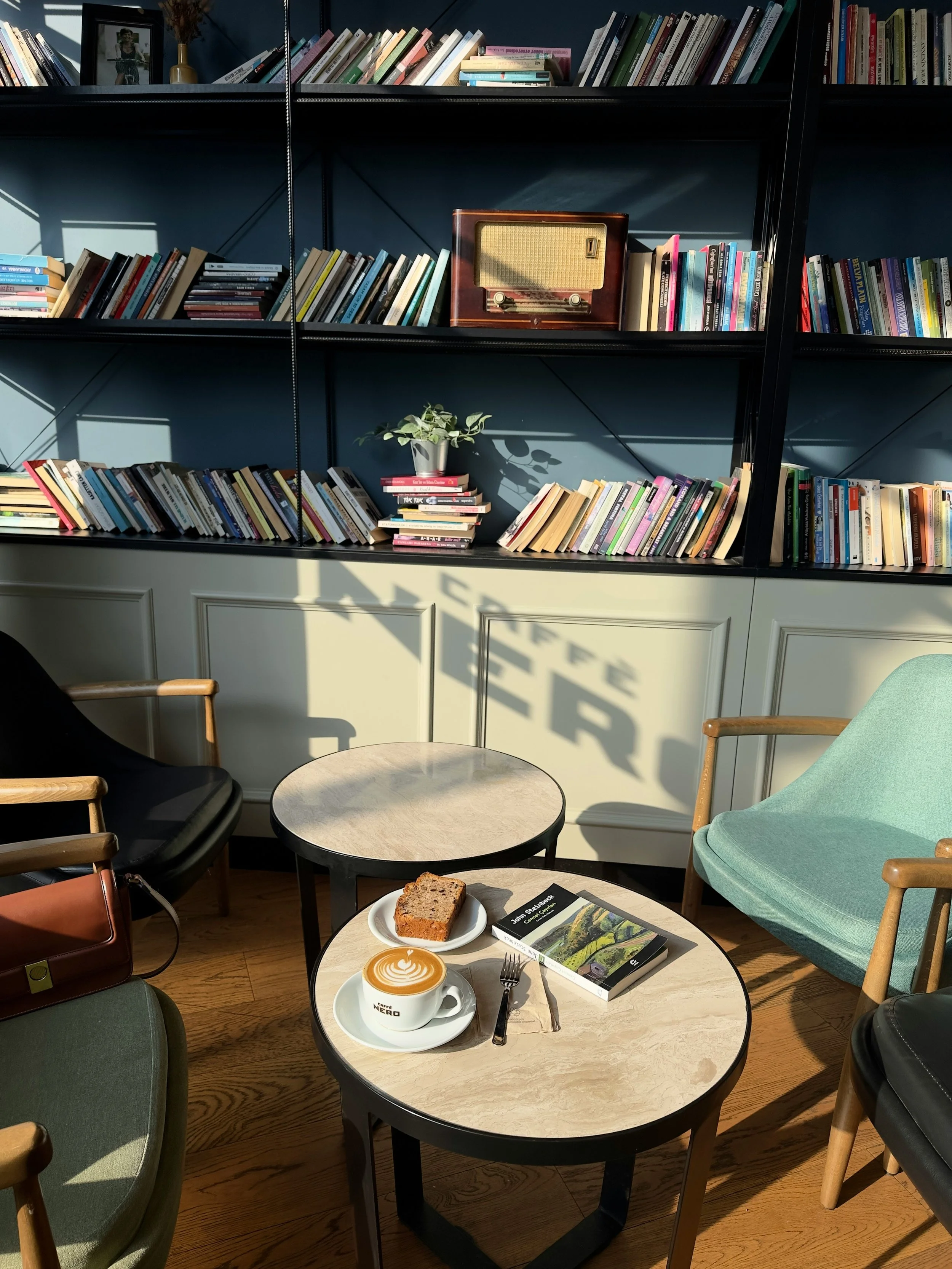Interior of a cozy café with a round table holding a coffee with latte art, a slice of cake, a book, and utensils. Surrounding chairs include a green upholstered armchair and wooden chairs. In the background, a black bookshelf filled with books and a vintage radio with a plant are visible, illuminated by natural sunlight.