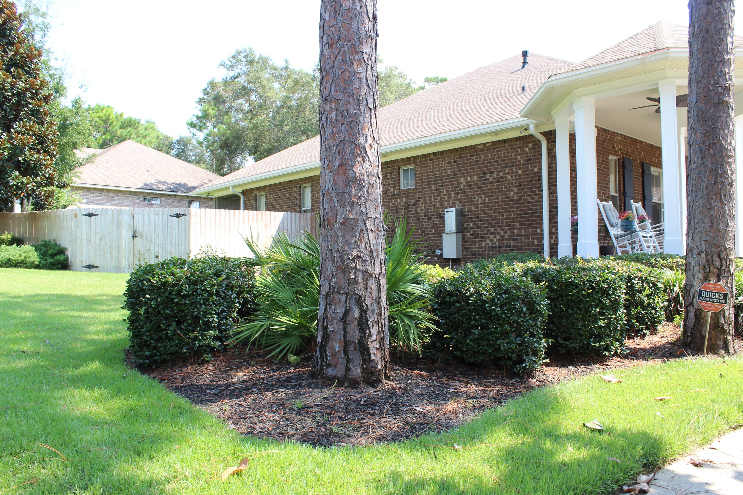 View of a suburban backyard with green lawn, bushes, trees, a brick house with a porch, rocking chairs with flowers, and a wooden fence with security alarm sign.
