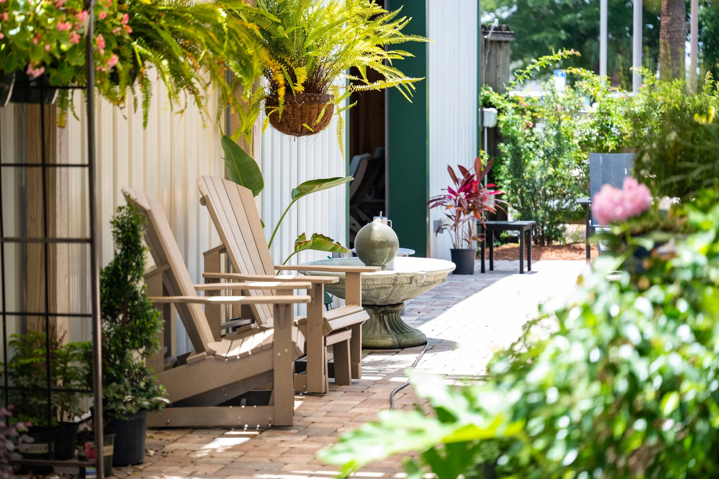 A backyard patio with wooden chairs, a stone table, and various potted plants and hanging greenery, with trees and a fence in the background.