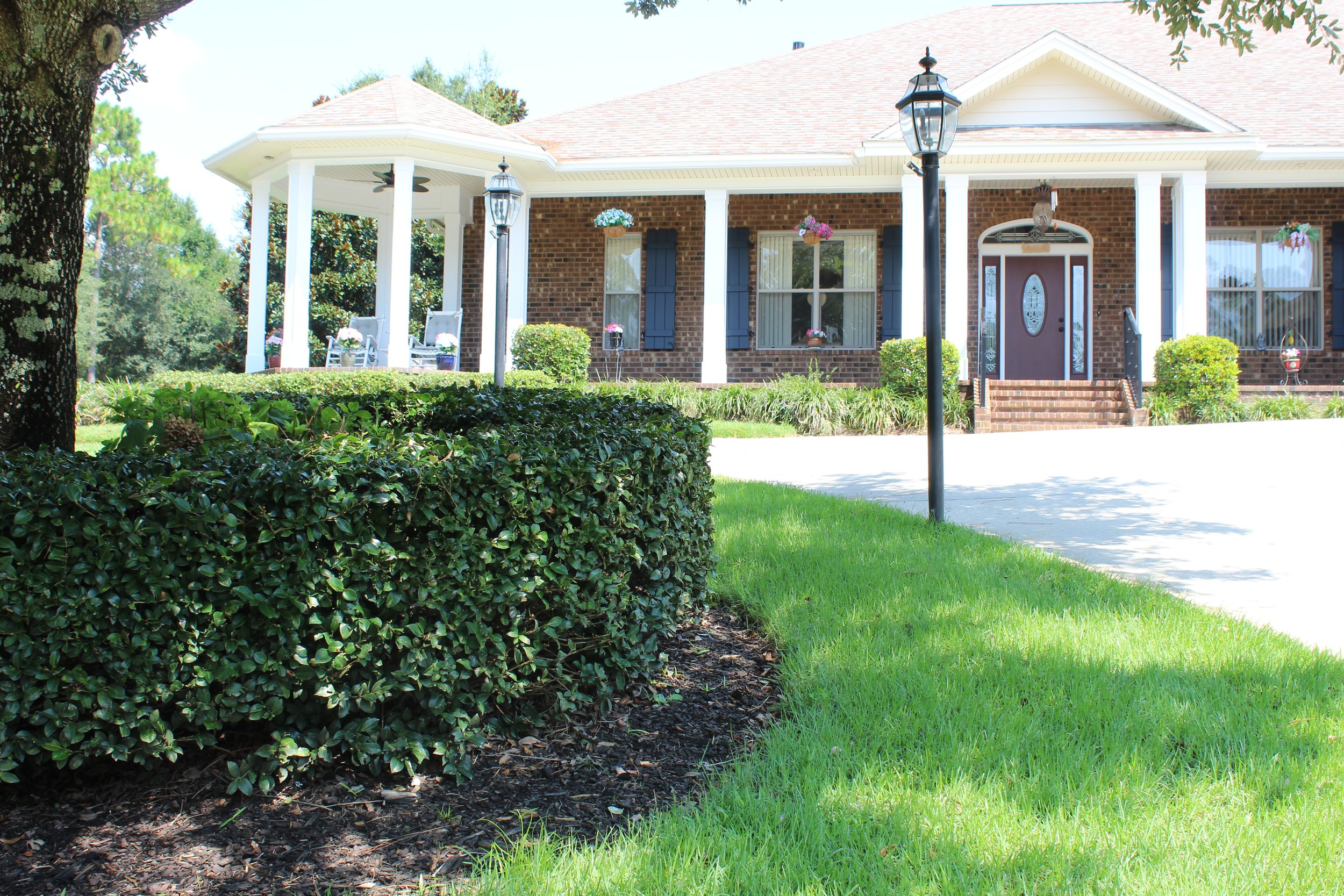Front view of a brick house with a porch, white columns, hanging flower baskets, a wheelchair ramp, and a landscaped yard with green grass, bushes, and trees.