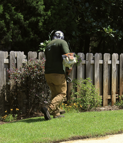 A man in a black helmet and dark green shirt using a leaf blower to clear leaves and debris from a backyard lawn.