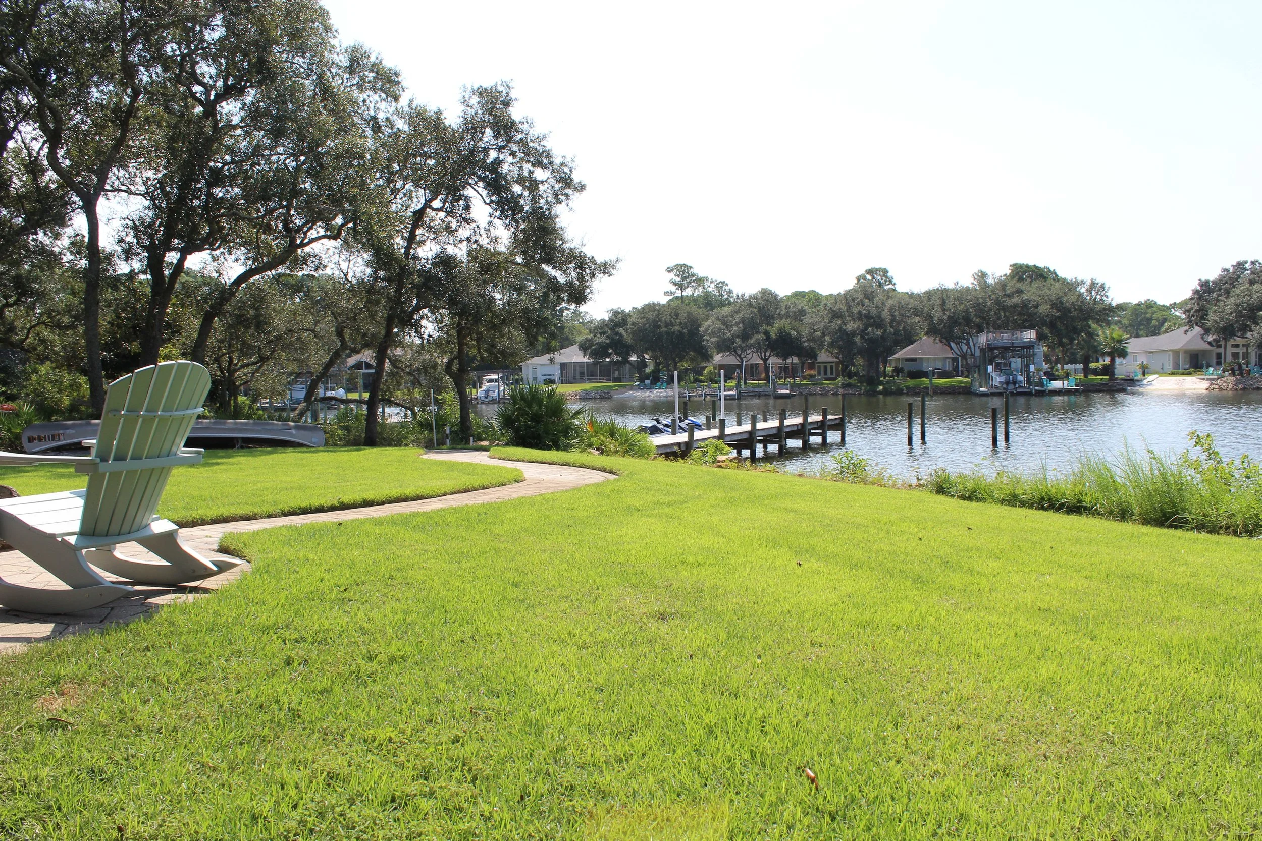 A lakeside backyard scene with green grass, an Adirondack chair, well-maintained trees, a dock leading to calm water, and houses along the shoreline.