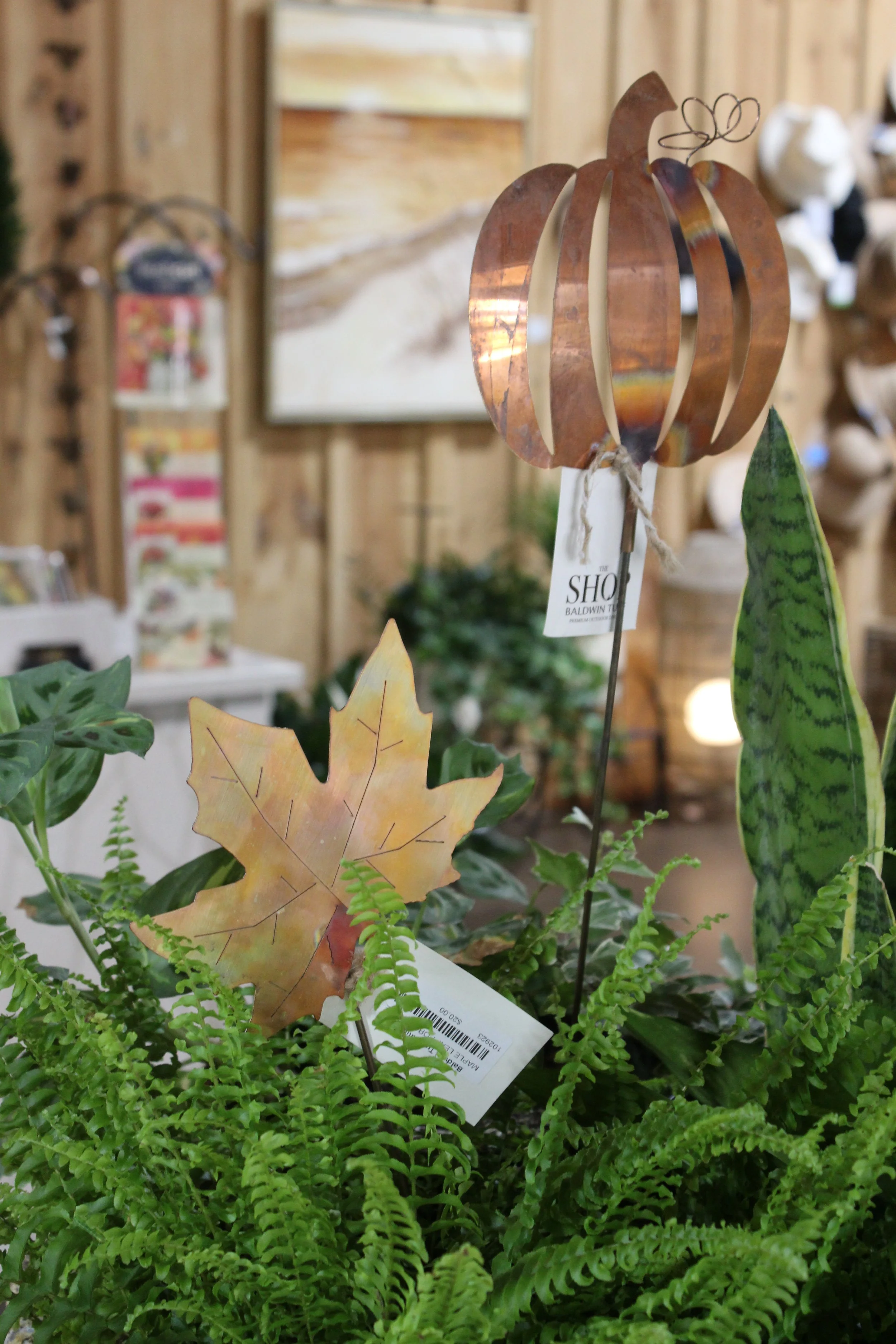 A decorative metal pumpkin sculpture surrounded by artificial leaves and green plants, with a blurred wooden wall and picture in the background.