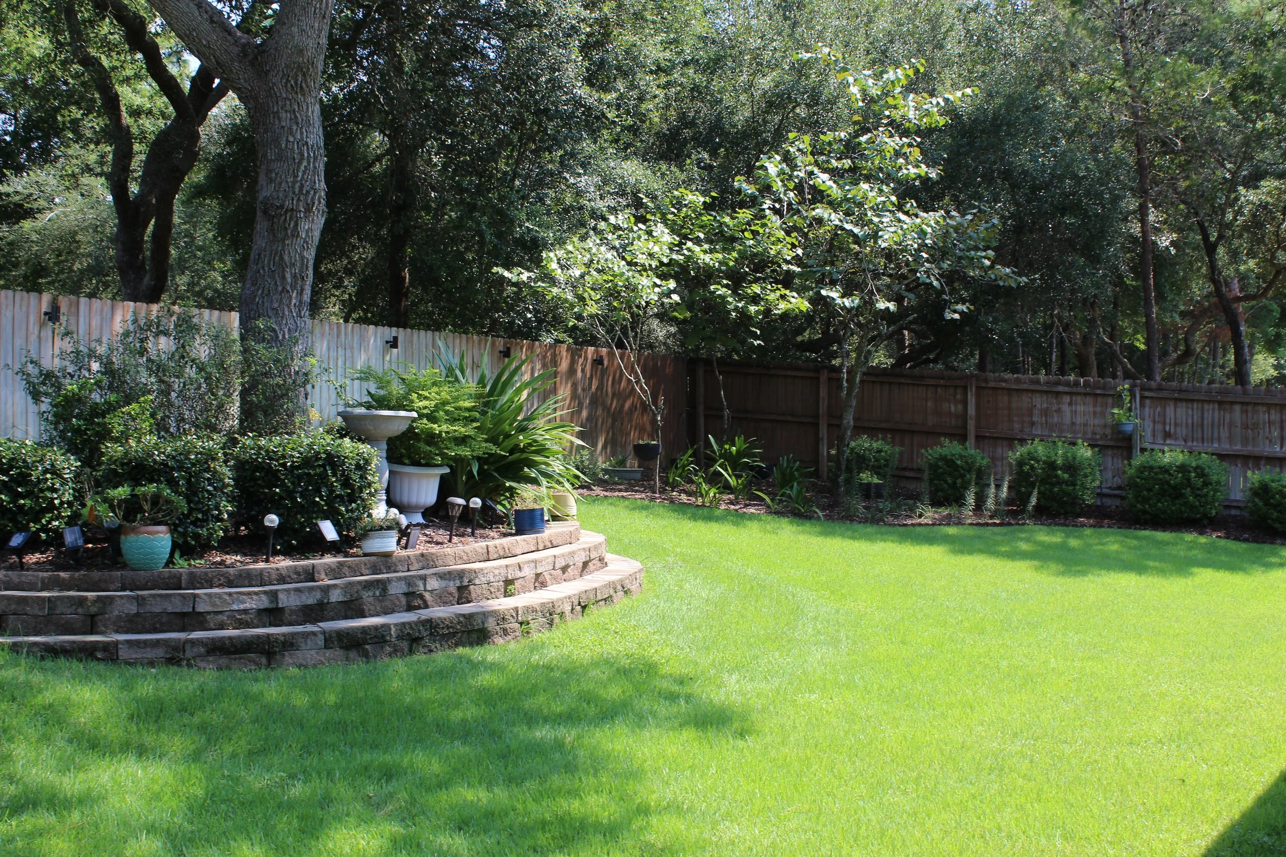 A backyard garden with a green lawn, potted plants, and a bordered flower bed. There are tall trees, a wooden fence, and a birdbath in the garden.