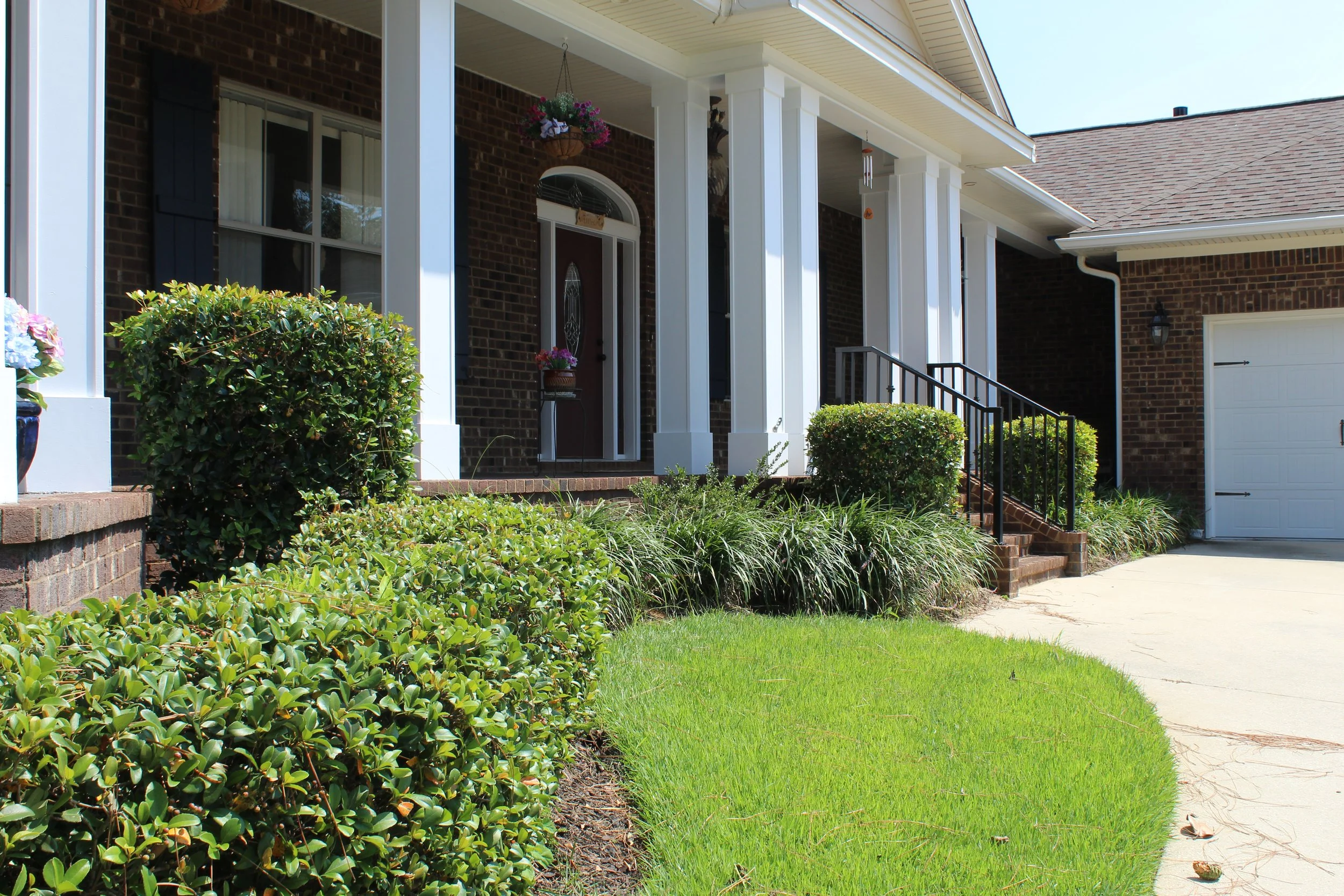 Front porch of a brick house with white columns, a red door with decorative glass, hanging flower basket, potted flowers, shrubbery, and stairs with black handrails leading to the entrance, beside a concrete sidewalk and garage with a white door.