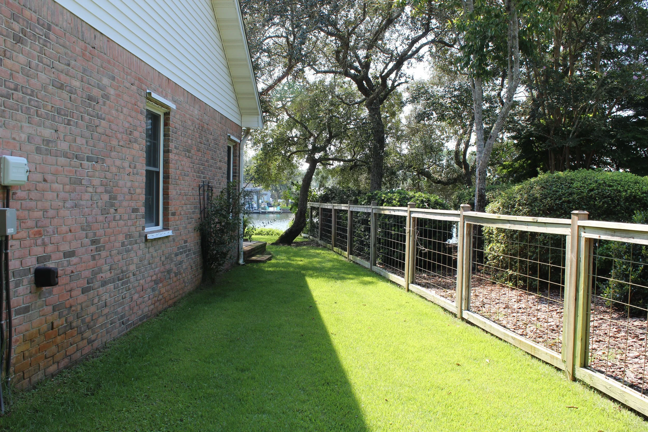 Side yard of a house with a brick wall, white siding, green grass, trees, and a fence near a waterway.