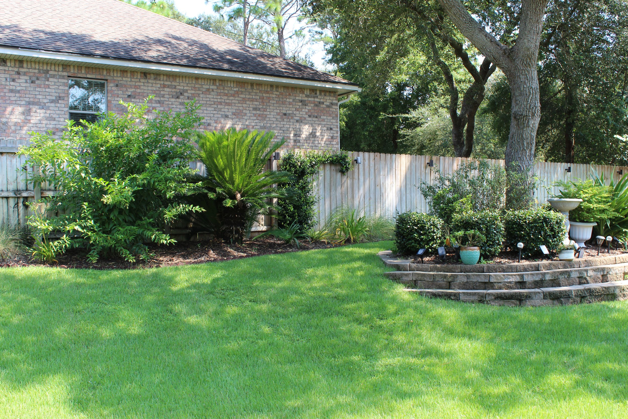 A well-maintained backyard with a lush green lawn, a variety of shrubs, and potted plants along a curved brick-bordered garden bed. There are two large trees, a wooden fence, and part of a brick house with a sloped roof in the background. The scene i