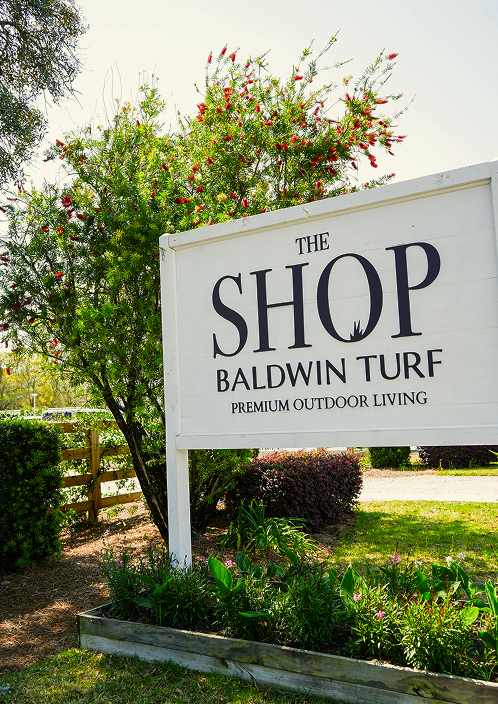 Sign reading "The Shop Baldwin Turf Premium Outdoor Living" in front of flowering shrub and trees in a landscaped outdoor setting.